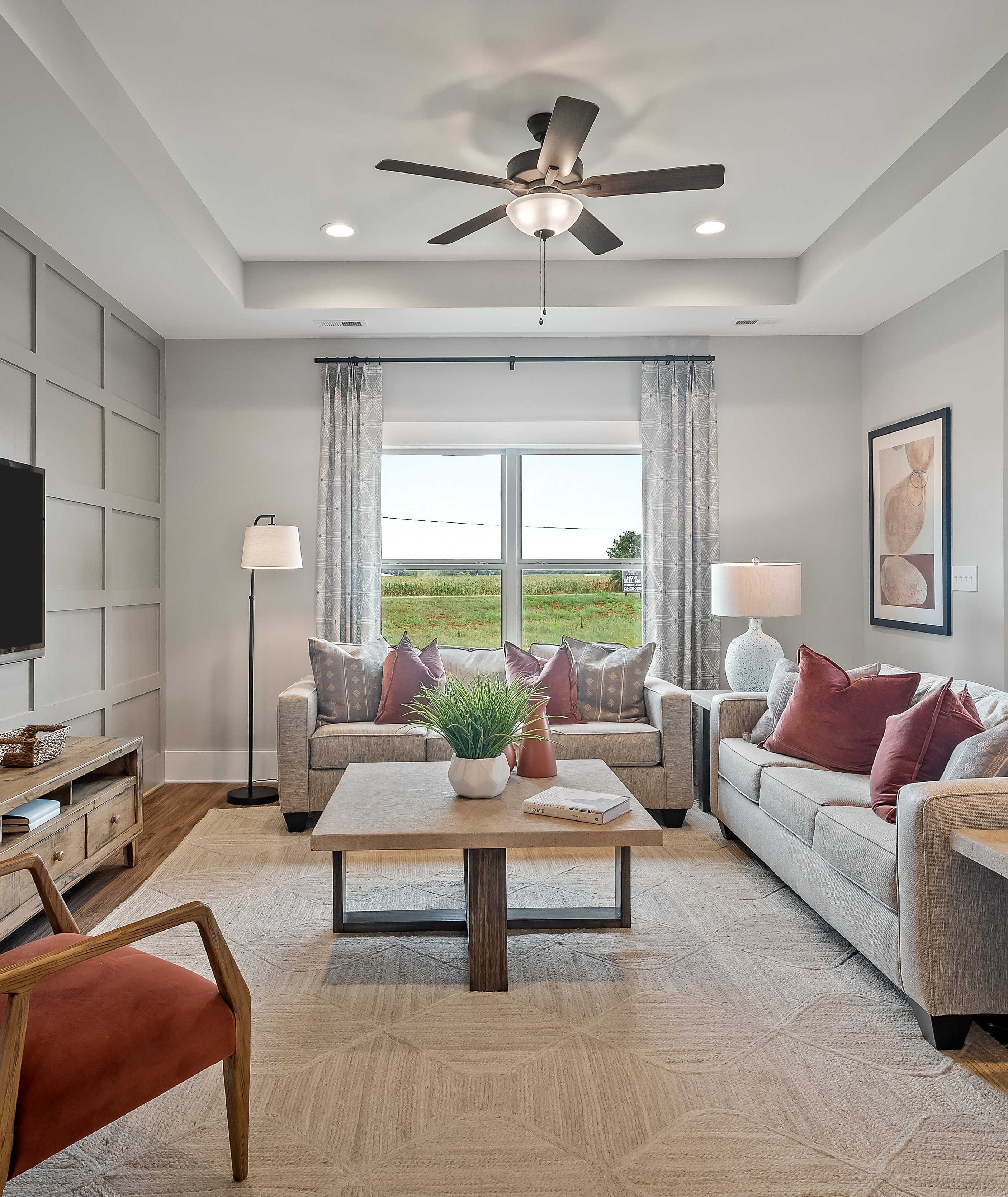 Spacious living room in Heritage Lakes home, New Market Alabama with gray shiplap walls, mounted TV, plush L-shaped sofa, ceiling fan, and scenic window views
