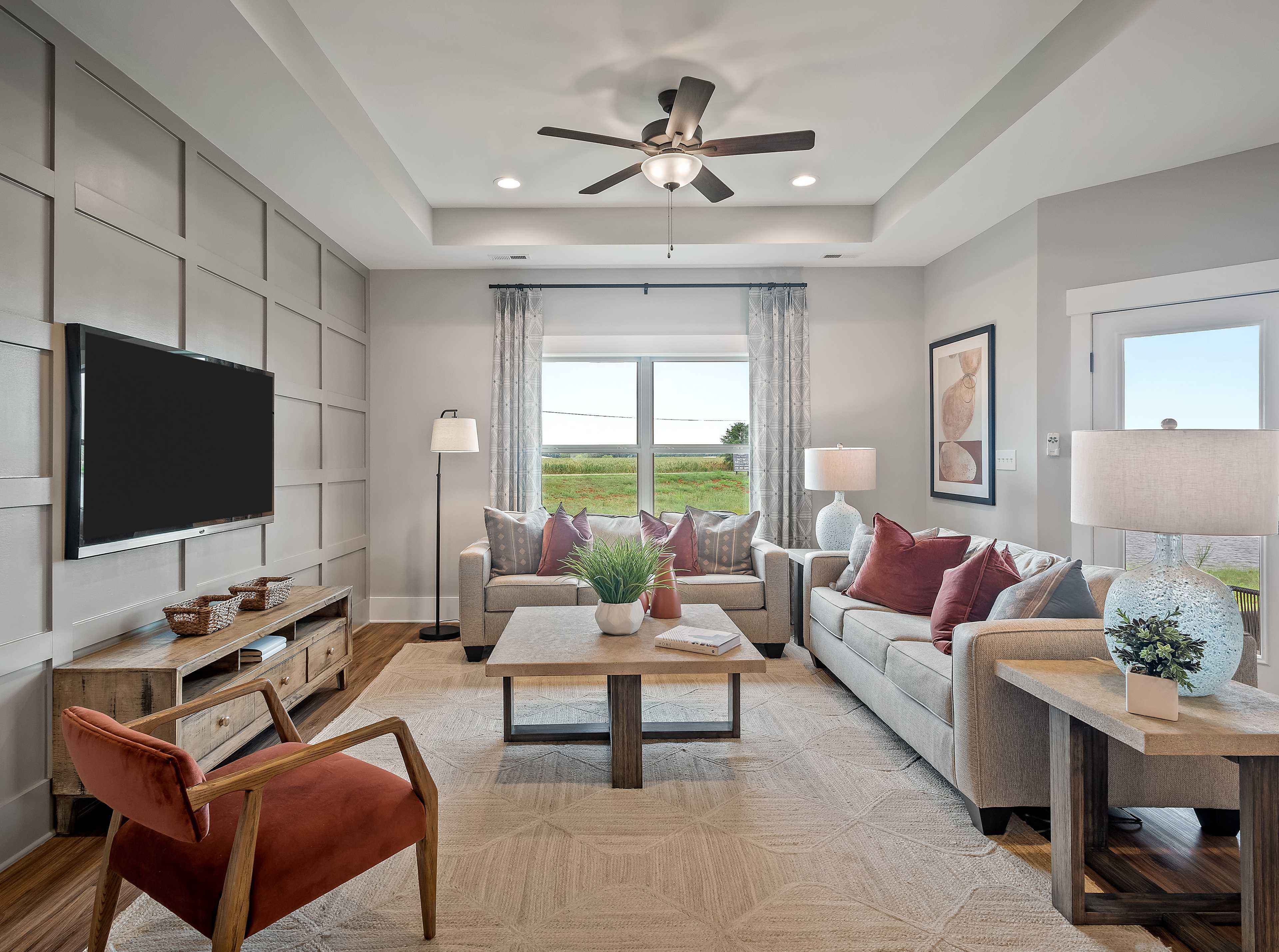 Spacious living room in Heritage Lakes home, New Market Alabama with gray shiplap walls, mounted TV, plush L-shaped sofa, ceiling fan, and scenic window views