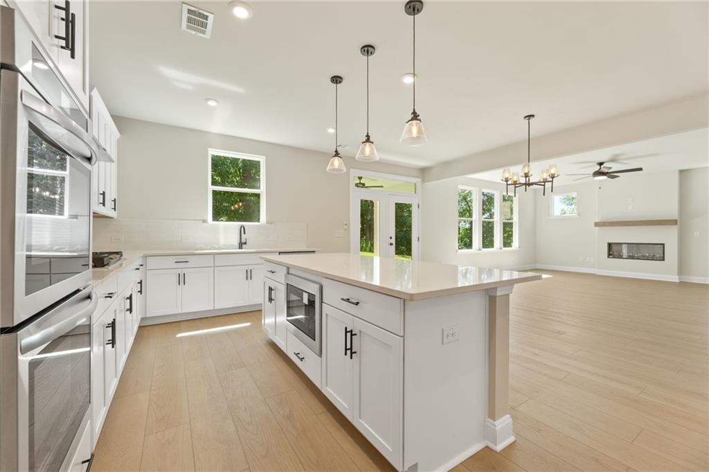 Modern open-concept kitchen with white cabinets, large island, stainless double ovens, pendant lights, and adjacent living room fireplace in The Hickory E, Buford, GA