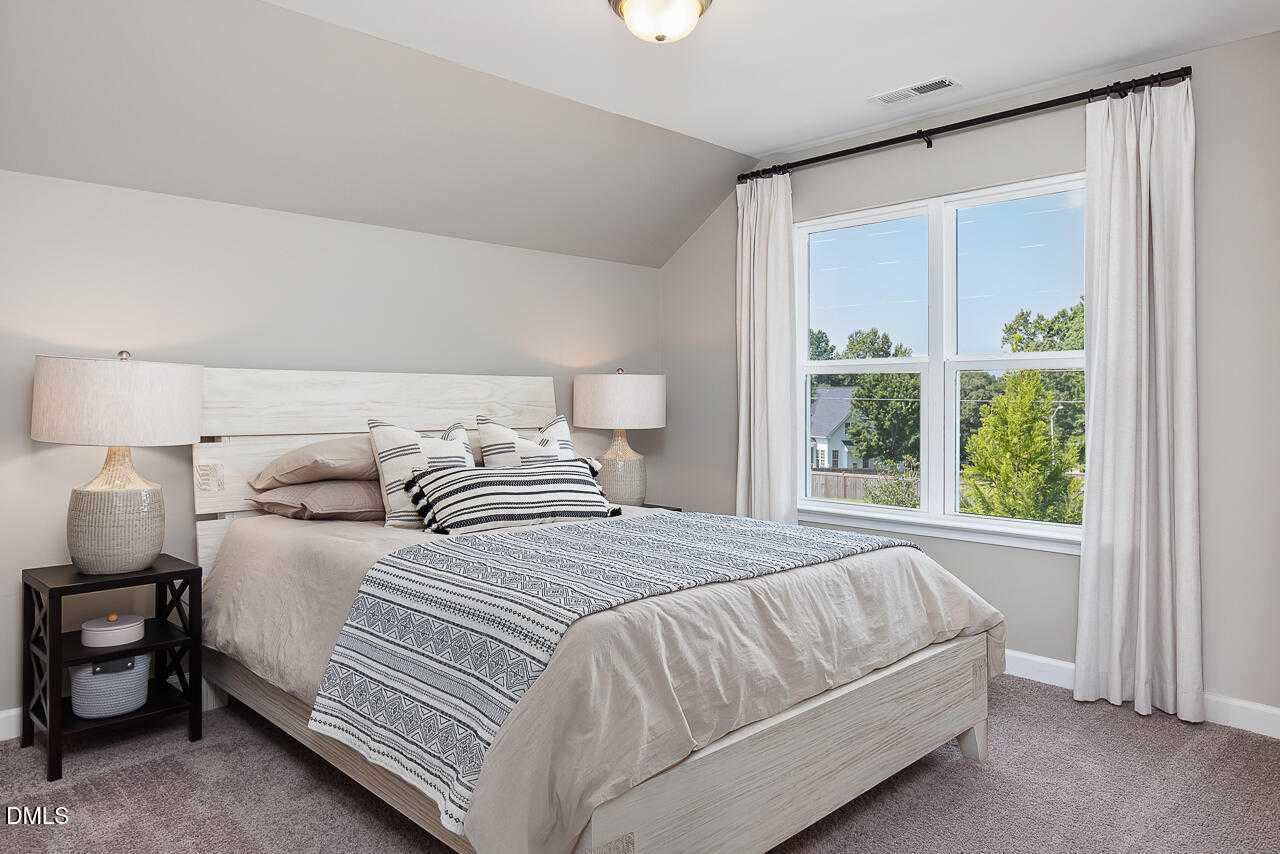 Bright bedroom with white wooden bed, neutral bedding, and large window view of trees in Davidson Homes The Ash B, Lillington, NC