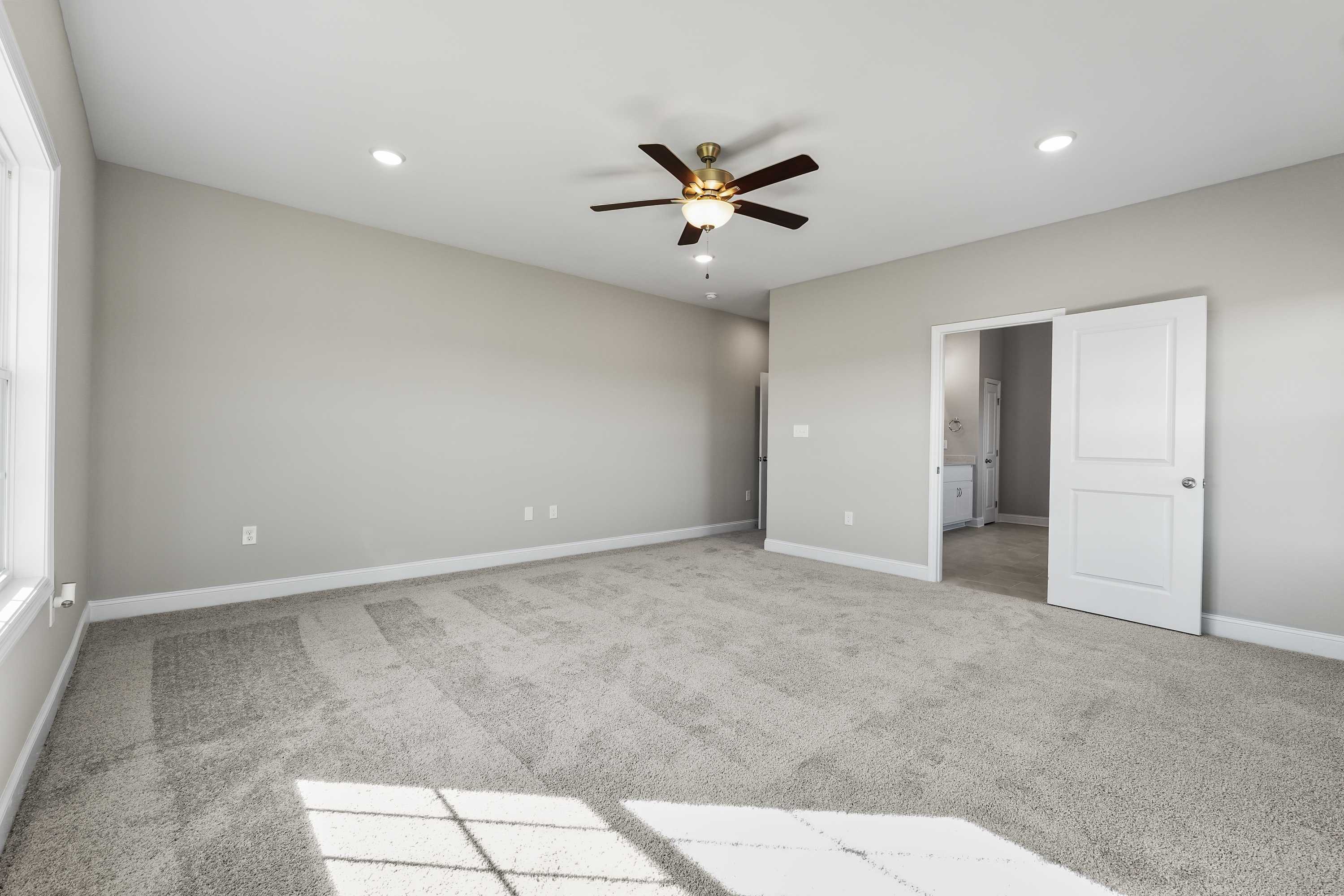 Spacious master bedroom in The Valencia home design with neutral walls, ceiling fan, and adjacent bathroom entry