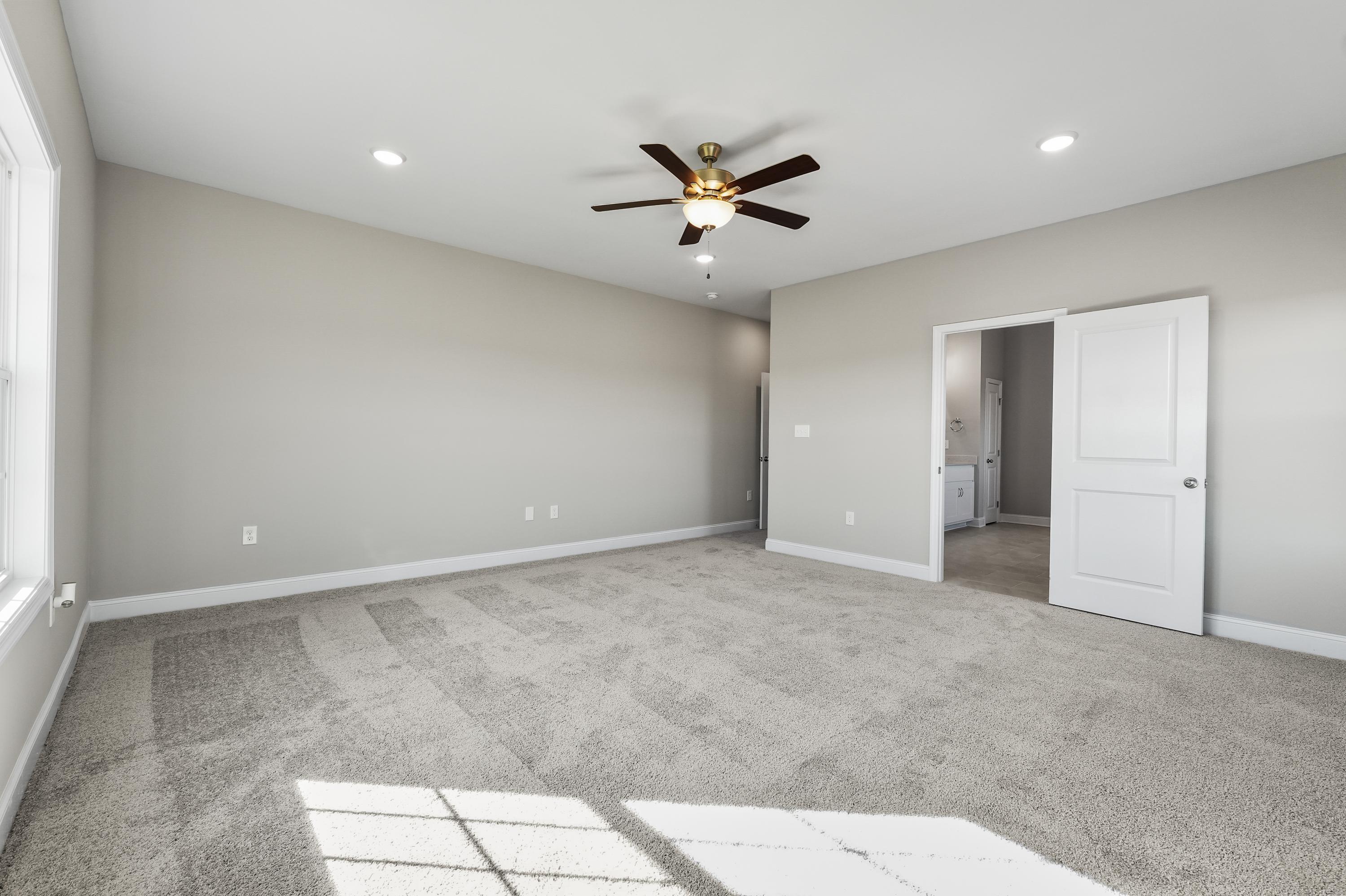Spacious master bedroom in The Valencia home design with neutral walls, ceiling fan, and adjacent bathroom entry
