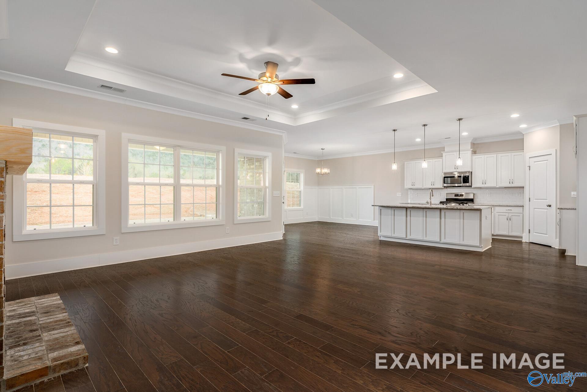 Open-concept great room with stone fireplace, large windows, ceiling fan, and adjacent white kitchen in Davidson Homes The Rockford, Harvest, AL