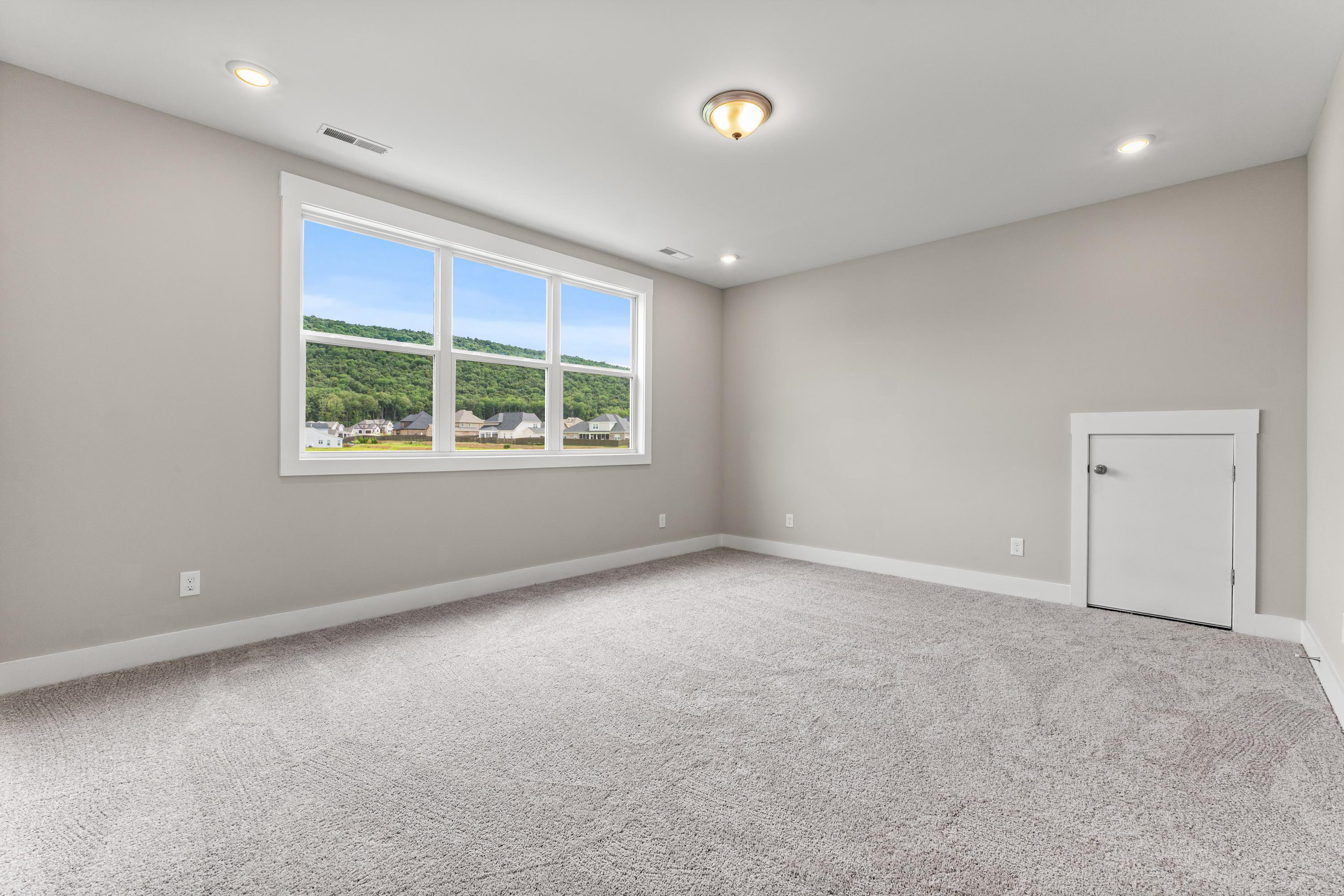 Spacious empty bedroom in The Oxford C with light gray walls, beige carpet, large triple window overlooking green landscape, and recessed lighting