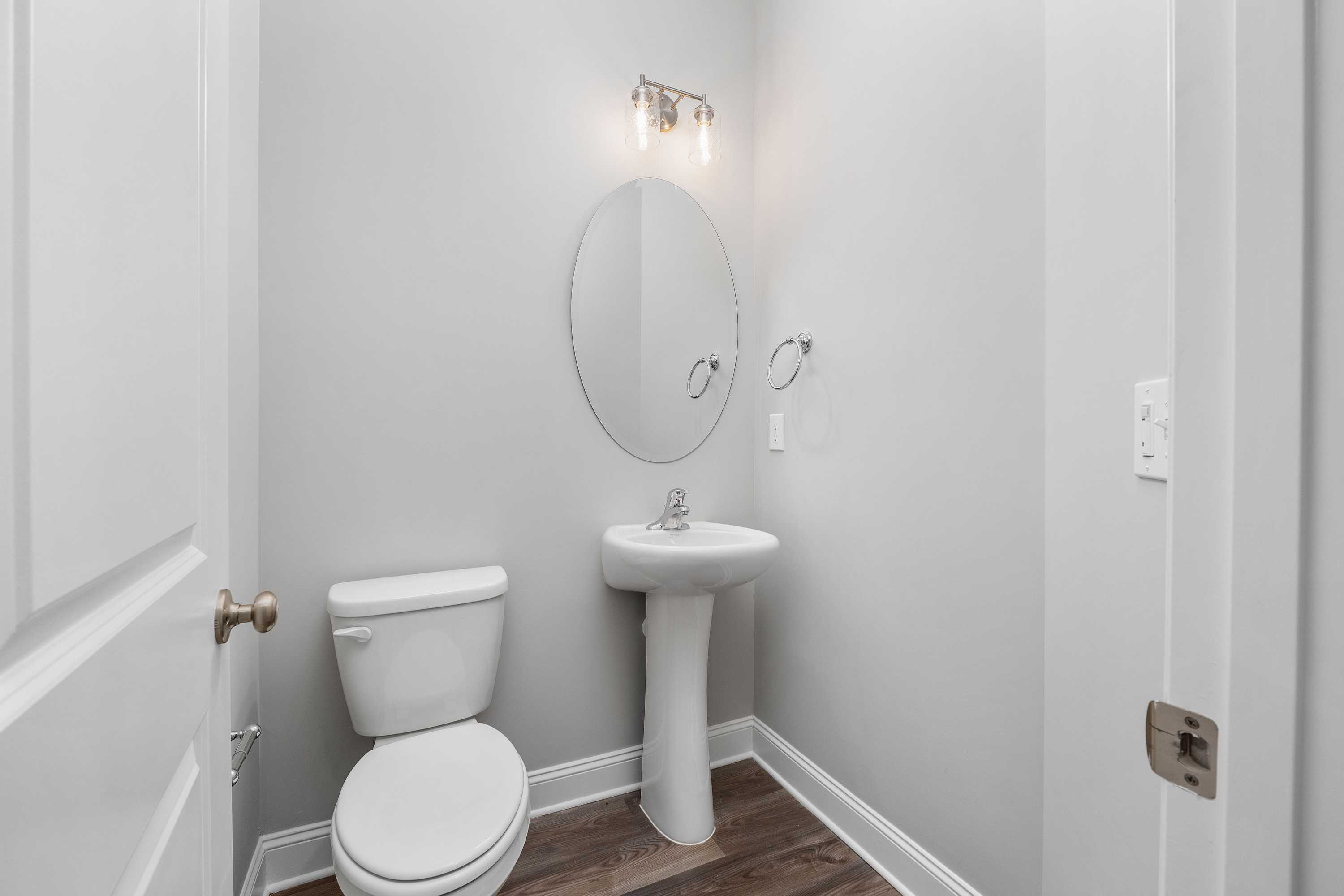 Cozy powder room in The Aiken home design featuring pedestal sink, round mirror, white toilet, and hardwood floors