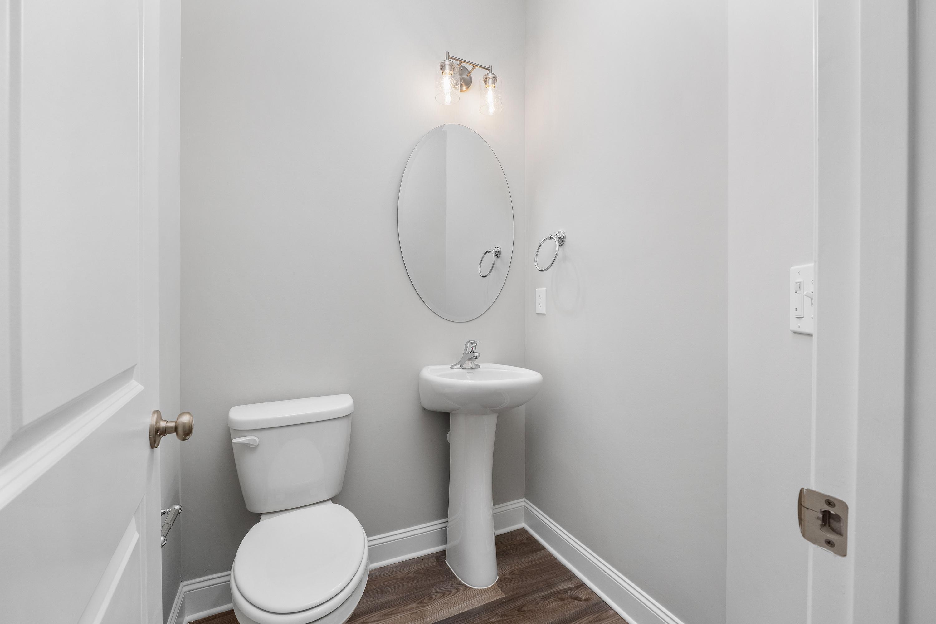 Cozy powder room in The Aiken home design featuring pedestal sink, round mirror, white toilet, and hardwood floors