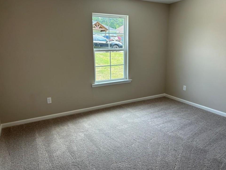 Bright empty bedroom with beige walls, carpeted floor, and large window overlooking green lawn in Davidson Homes The Washington, Phenix City