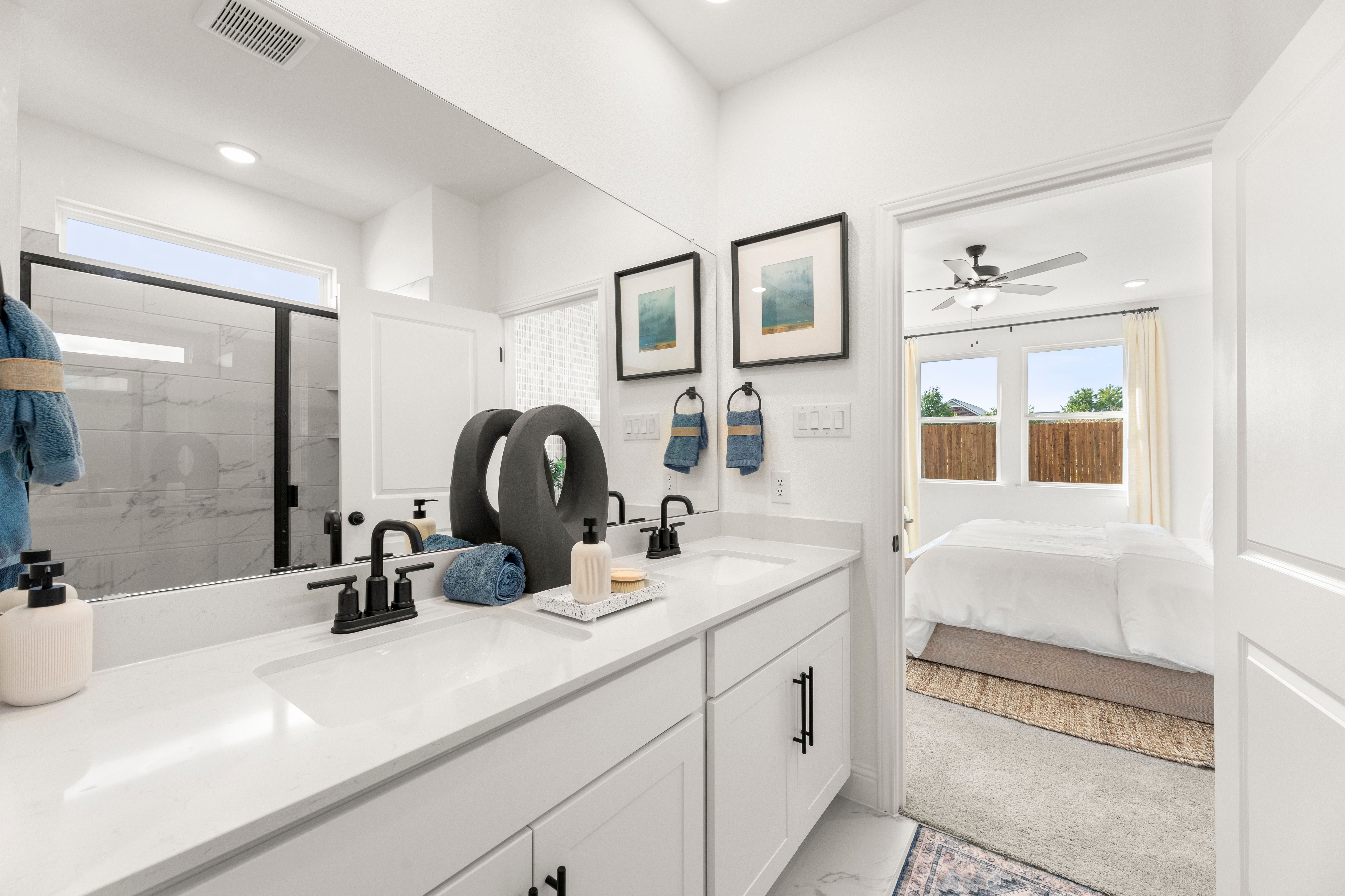 Spacious master bathroom in The Laurel featuring white double vanity, black faucets, walk-in shower, and bedroom view