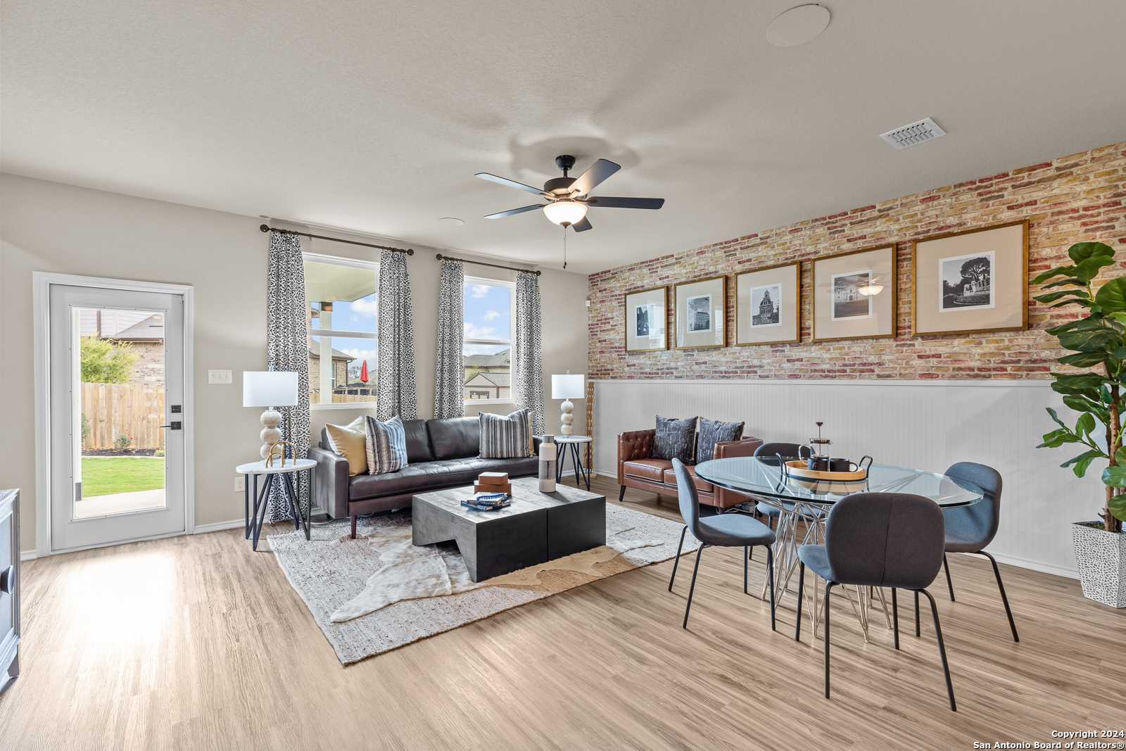 Modern living room with brick accent wall, gray sofas, round dining table, ceiling fan, and backyard view in Davidson Homes The Douglas F, Bricewood, San Antonio