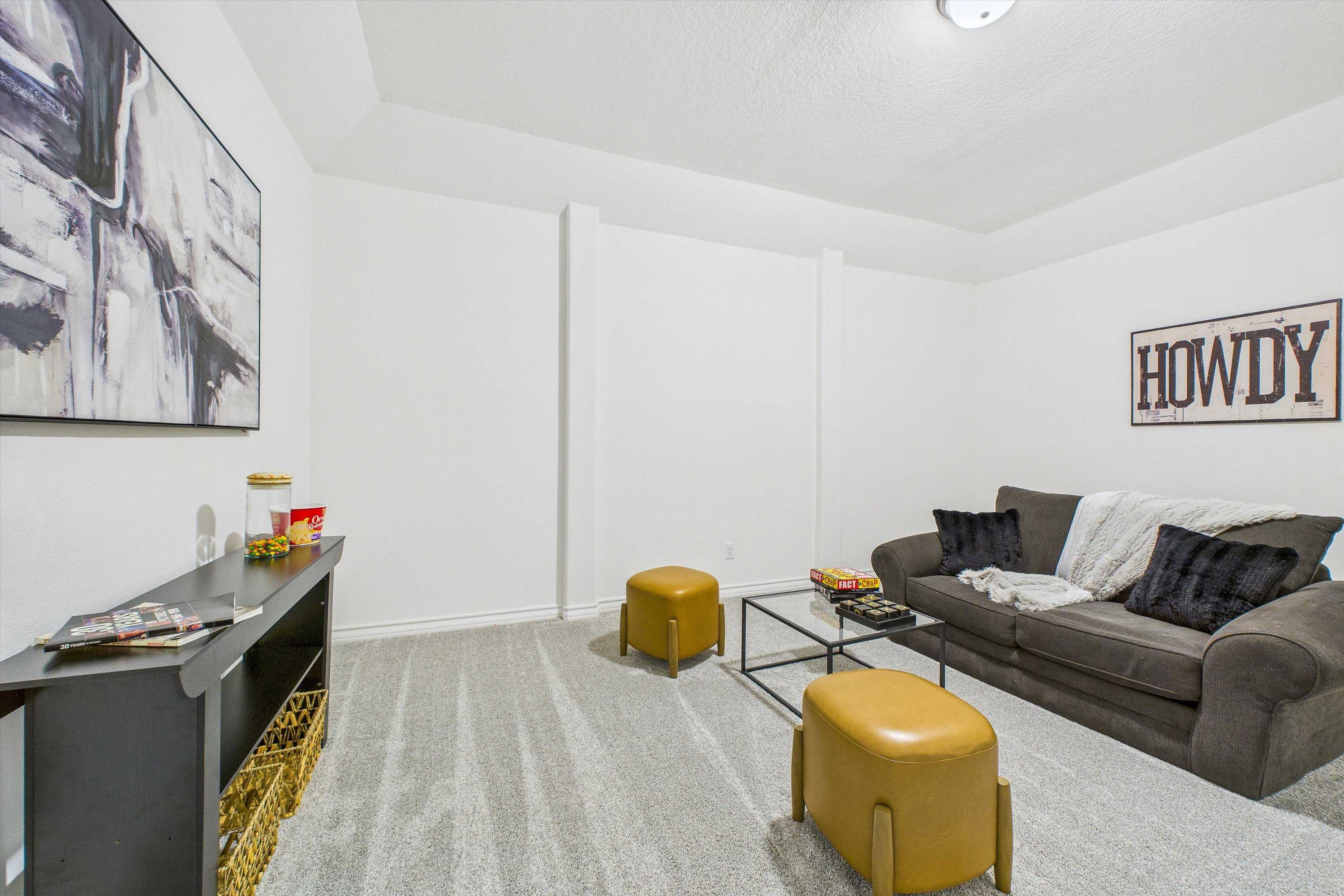 Cozy media room with gray sectional sofa, yellow poufs, abstract wall art, and Howdy sign in Davidson Homes The Summerlin C, Castroville TX
