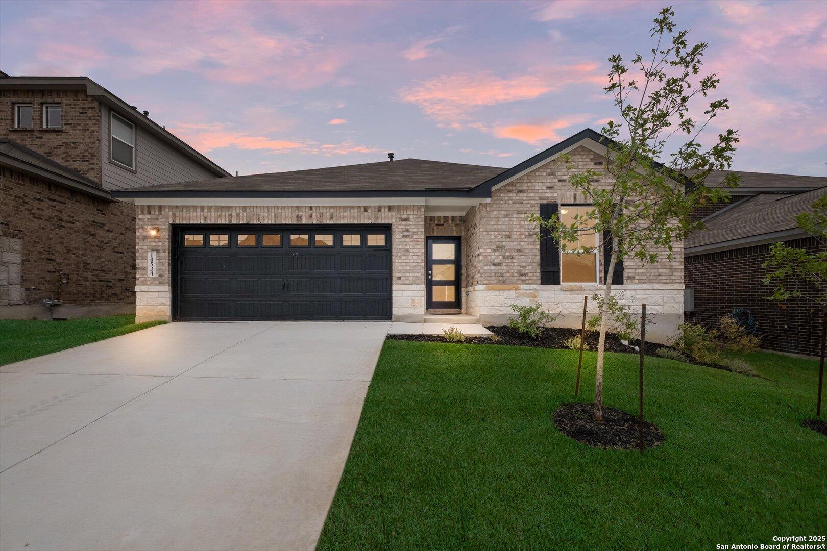 Modern brick single-story home with black 2-car garage, driveway, lush lawn, and young tree at sunset in Bricewood, San Antonio, Texas - Asheville K by Davidson Homes