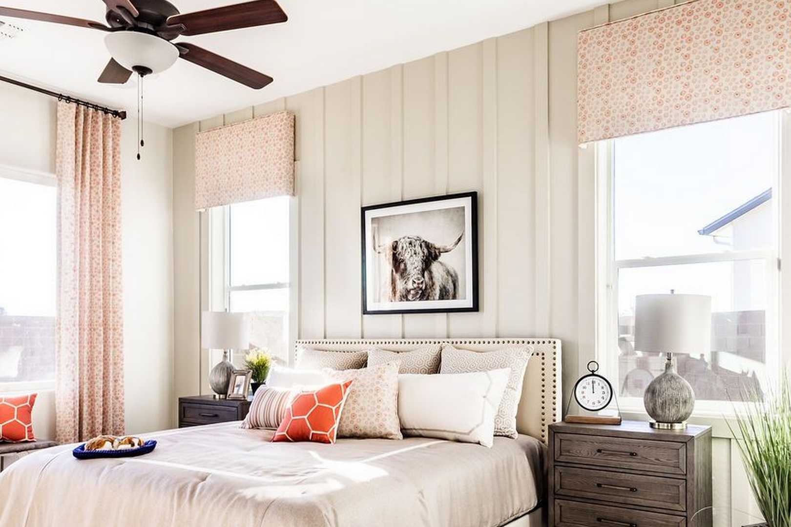 Spacious master bedroom in The Monarch A featuring king bed with orange accents, beige walls, large windows with patterned curtains, ceiling fan, and cow artwork