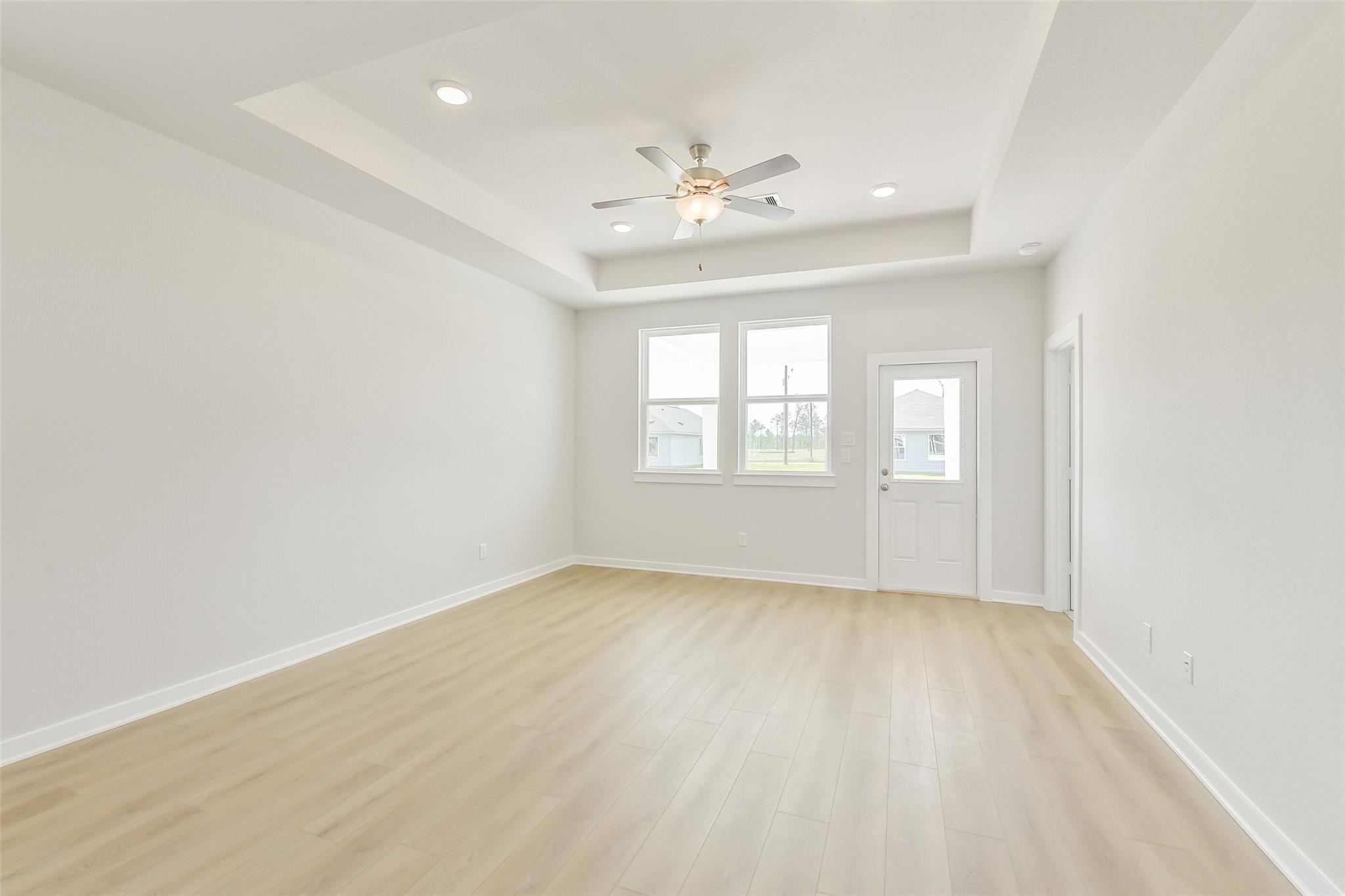 Bright living room with tray ceiling, ceiling fan, large windows, and light wood floors in Davidson Homes The Frio G, Conroe, Texas