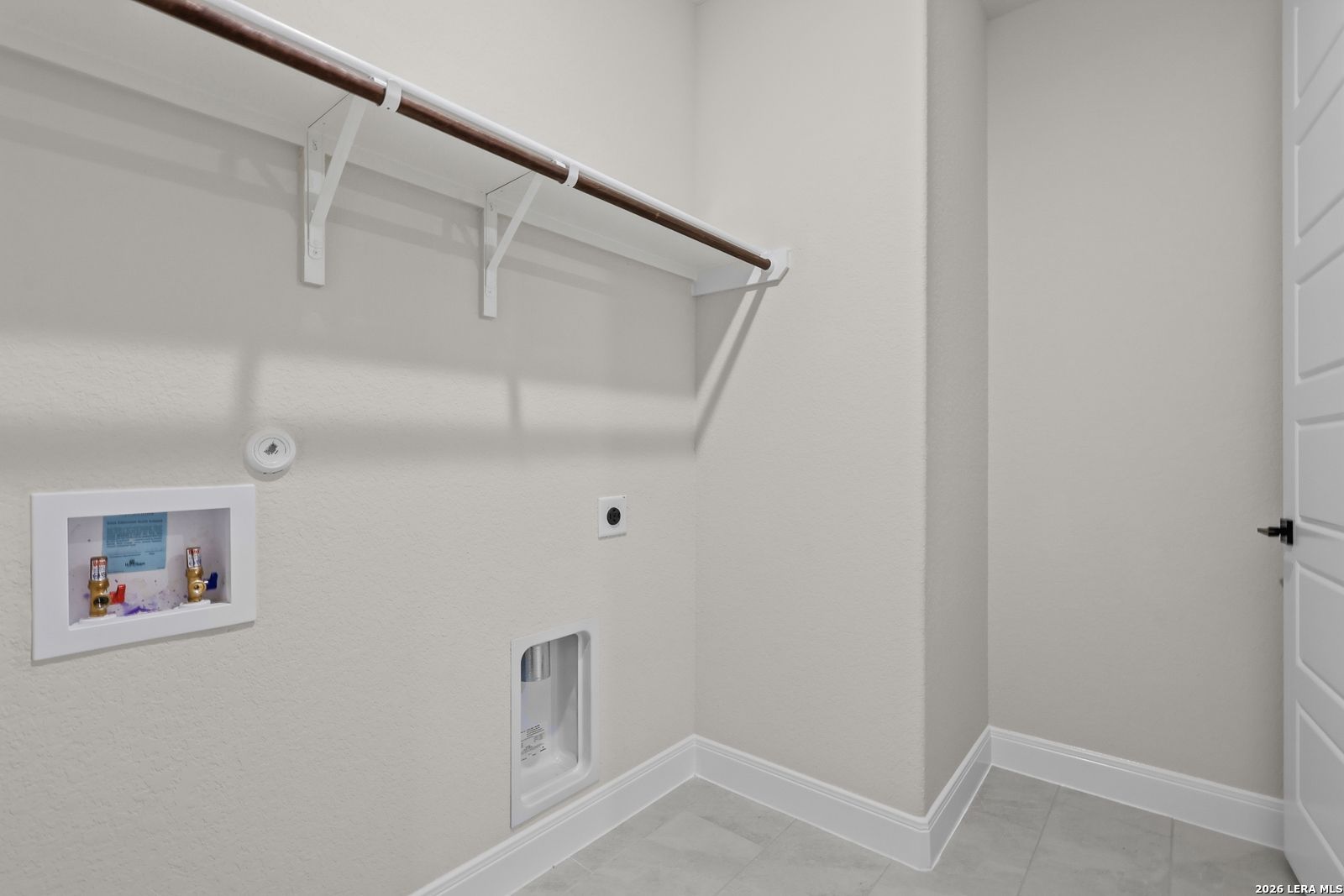 Functional laundry room with utility sink, wooden hanging rod, shelf, and beige tile floor in Davidson Homes The Lanier G, Castroville, Texas