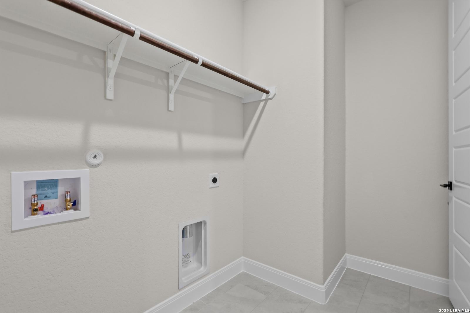 Functional laundry room with utility sink, wooden hanging rod, shelf, and beige tile floor in Davidson Homes The Lanier G, Castroville, Texas