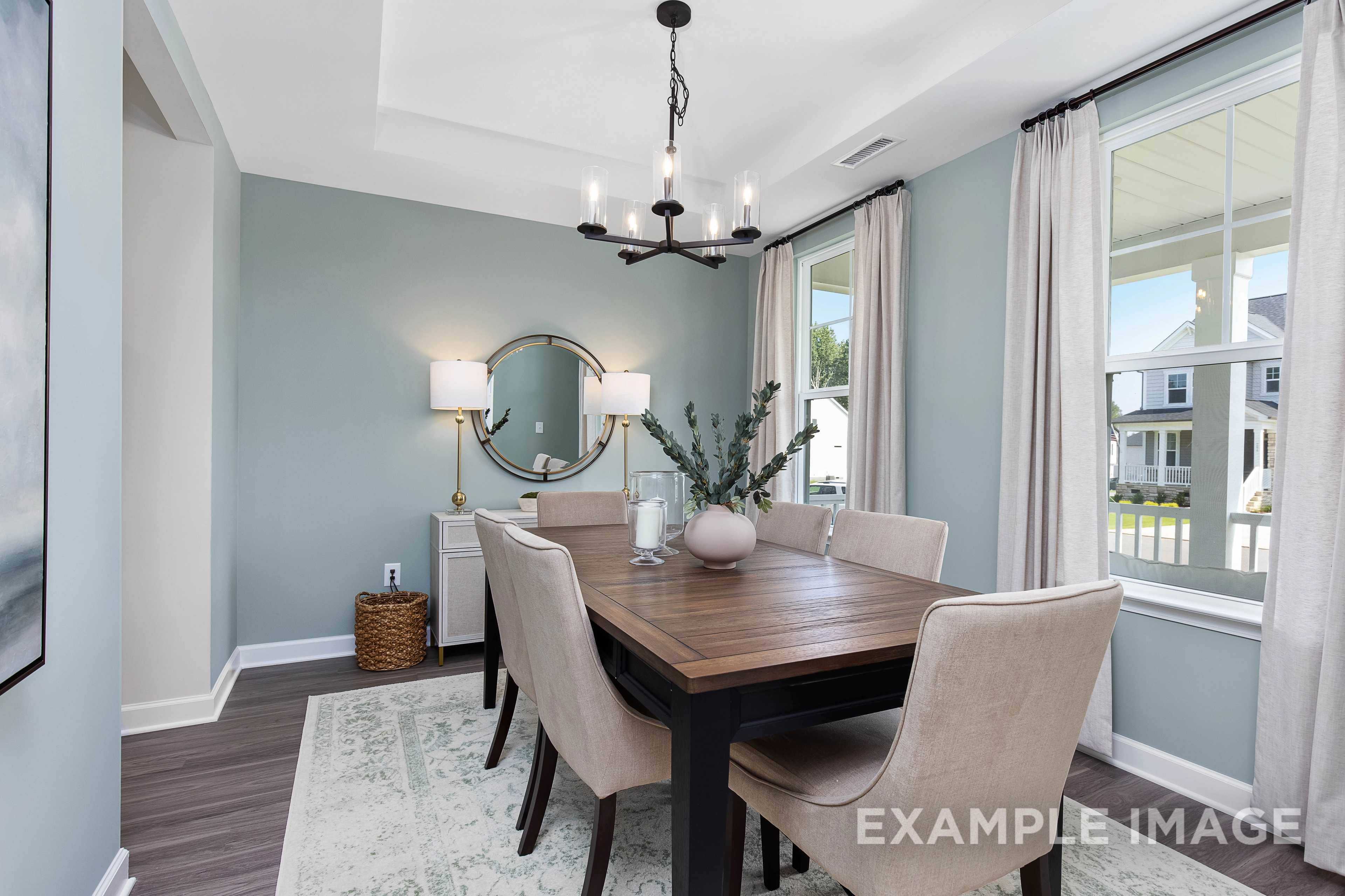 Cozy dining room in The Willow B with wooden farmhouse table, beige upholstered chairs, chandelier, round mirror, and large window