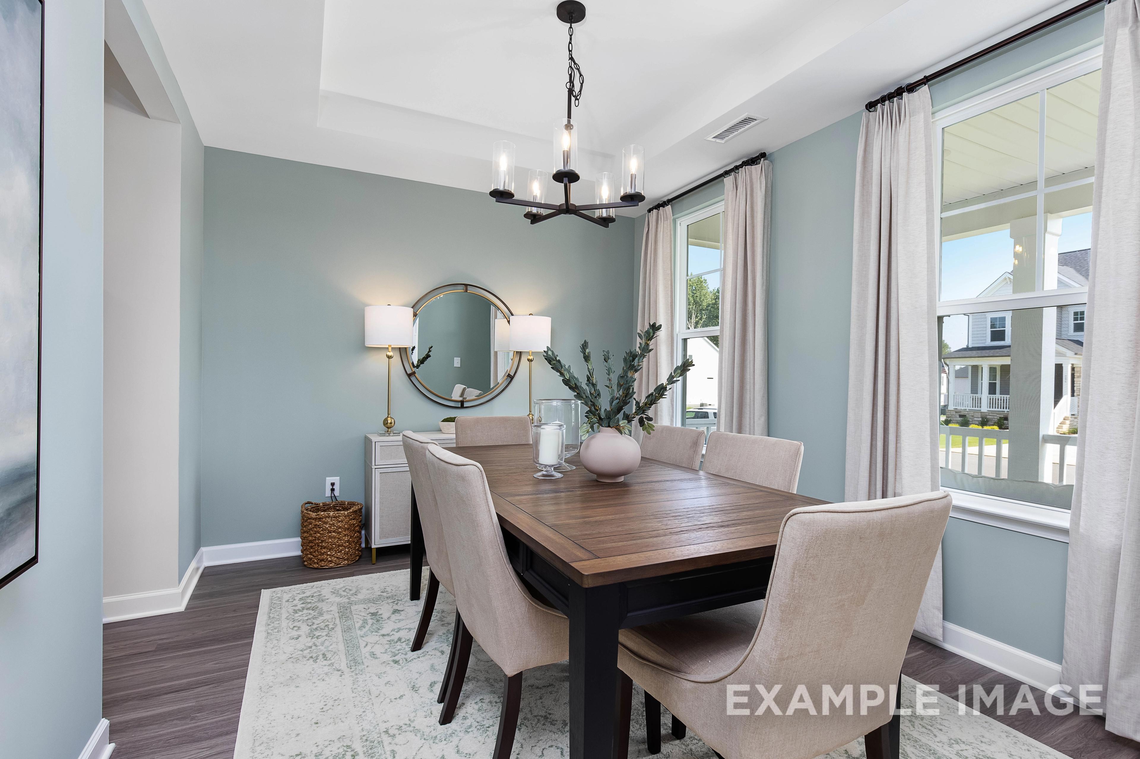 Cozy dining room in The Willow B with wooden farmhouse table, beige upholstered chairs, chandelier, round mirror, and large window