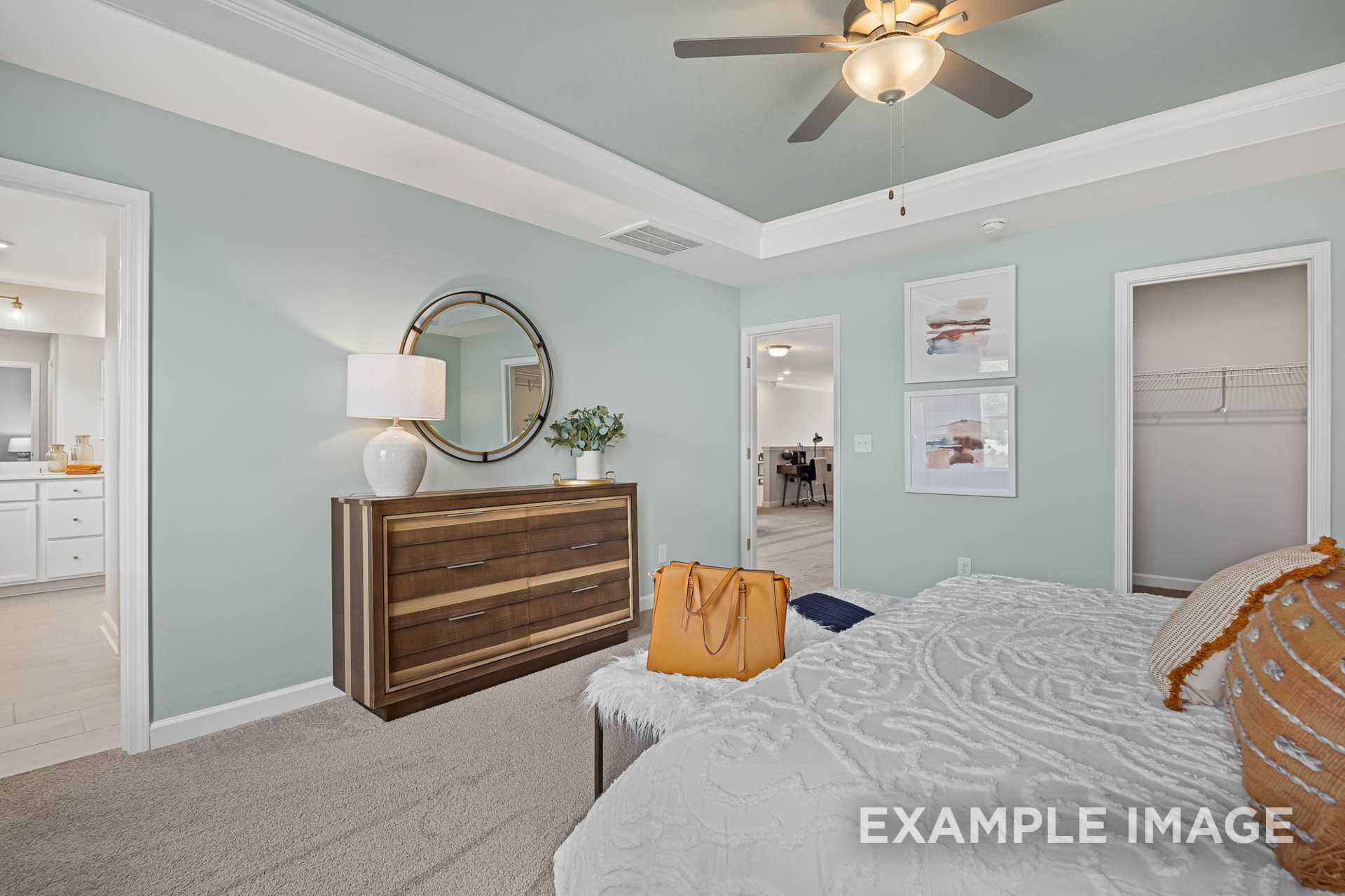 Spacious master bedroom in The Adalynn A with light blue walls, wooden dresser, round mirror, king bed, and ensuite access