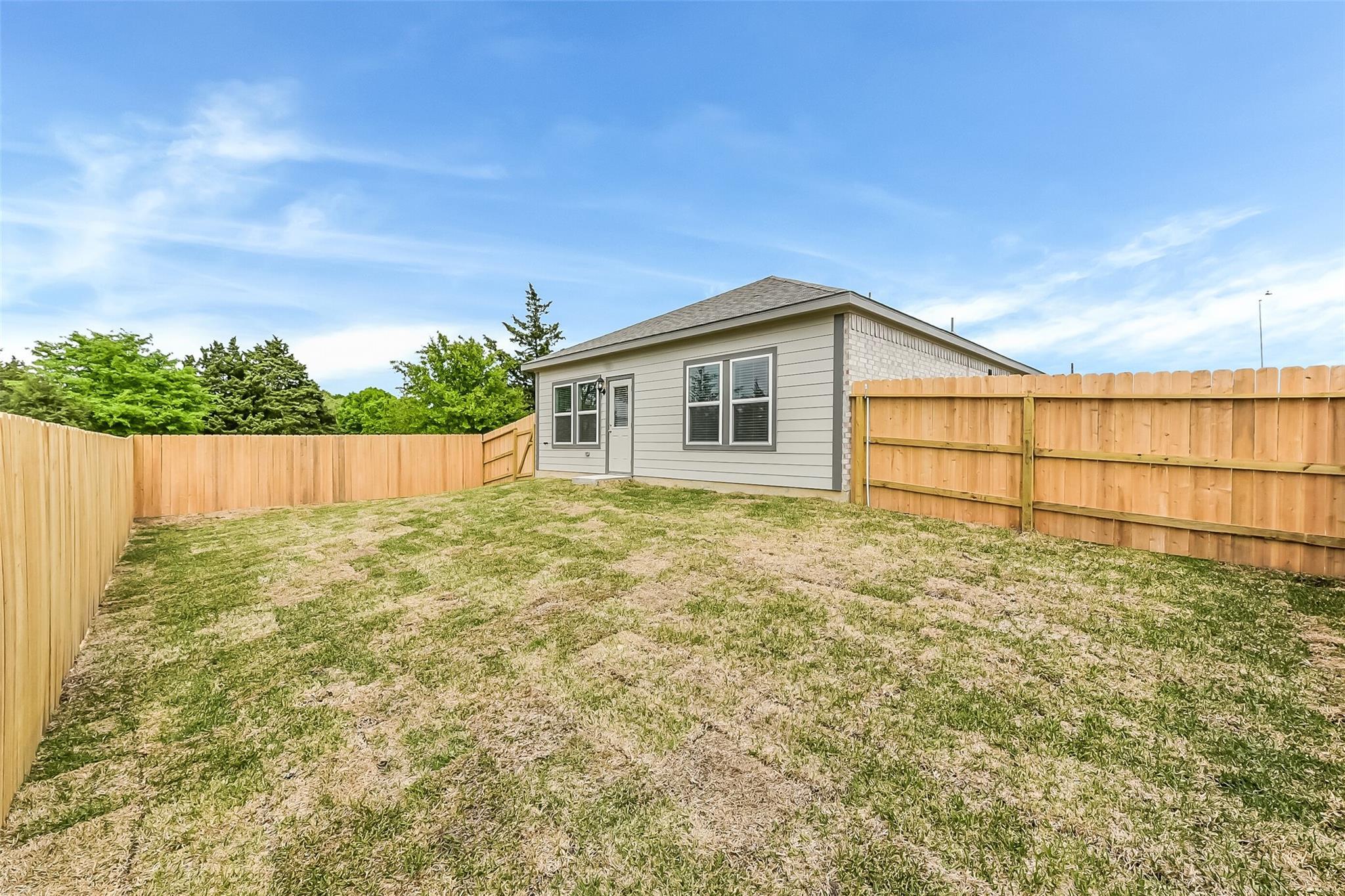 Single-story gray home with brick accents, wooden privacy fence, and grassy yard under blue sky in Windmill Estates, Magnolia, Texas