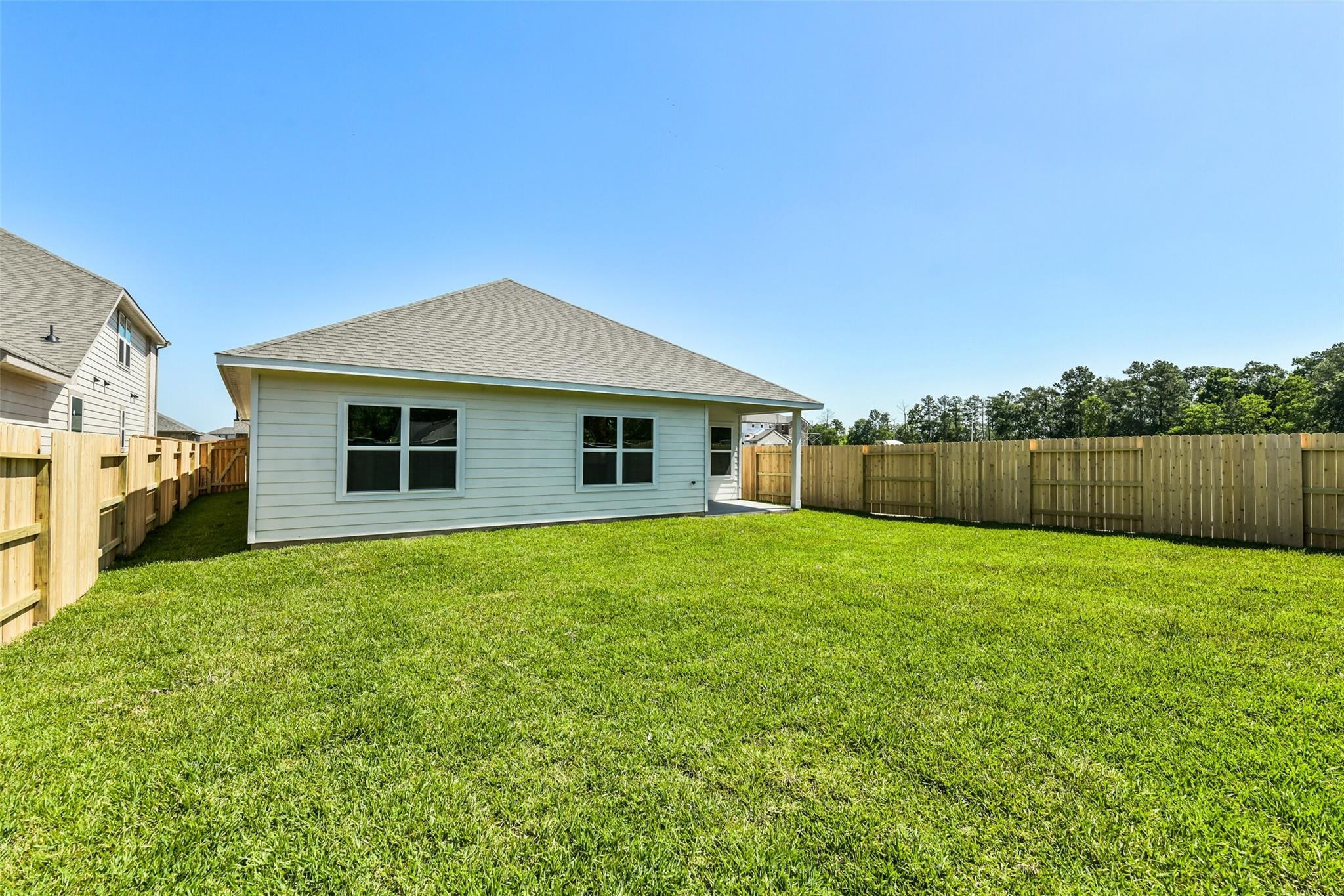 White single-story home side view with covered entry, double windows, and lush green fenced backyard in Sundance Cove, Crosby, Texas