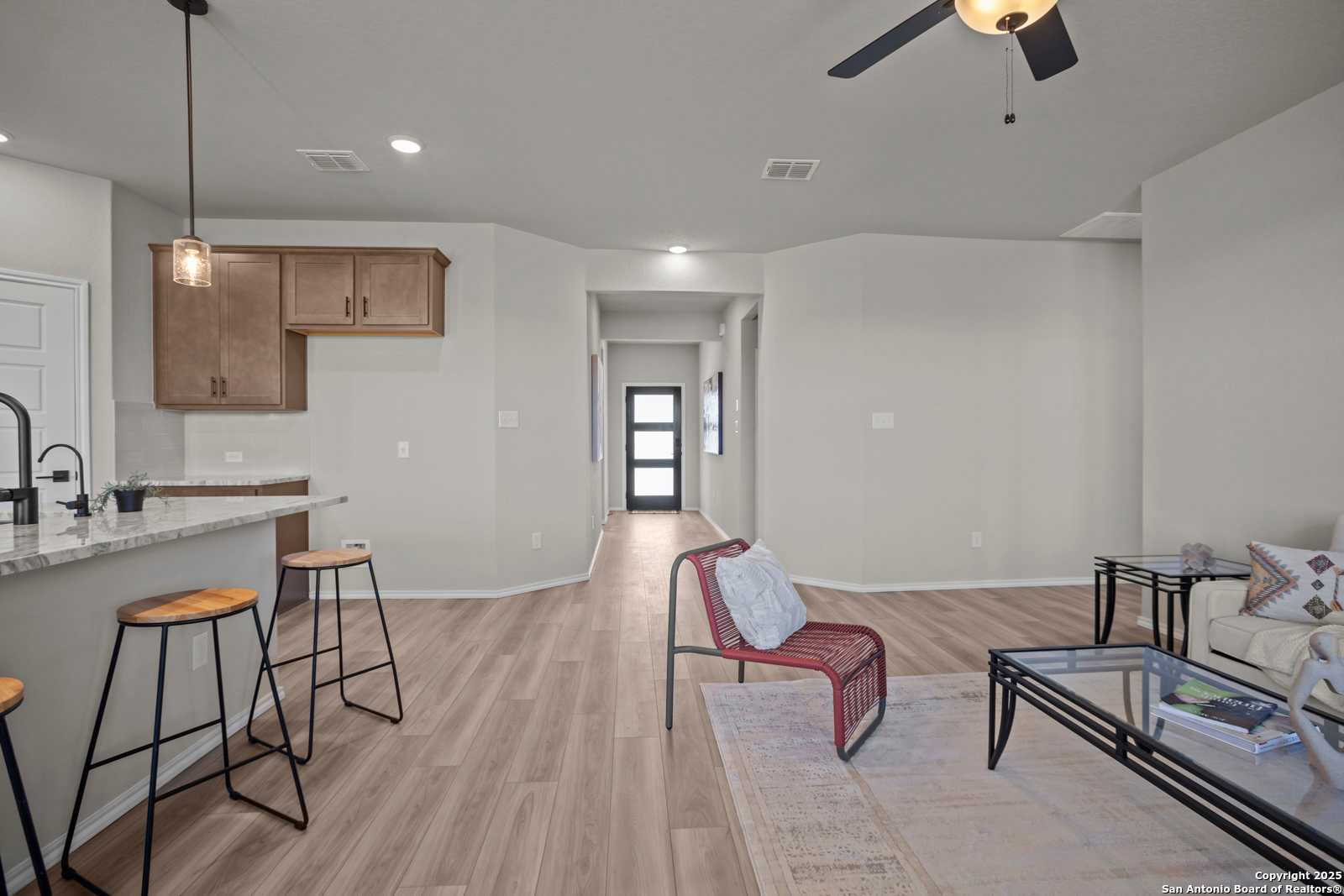 Open-concept living-kitchen in The Asheville H: wood cabinets, island with bar stools, ceiling fan, red accent chair, hardwood floors, Converse TX