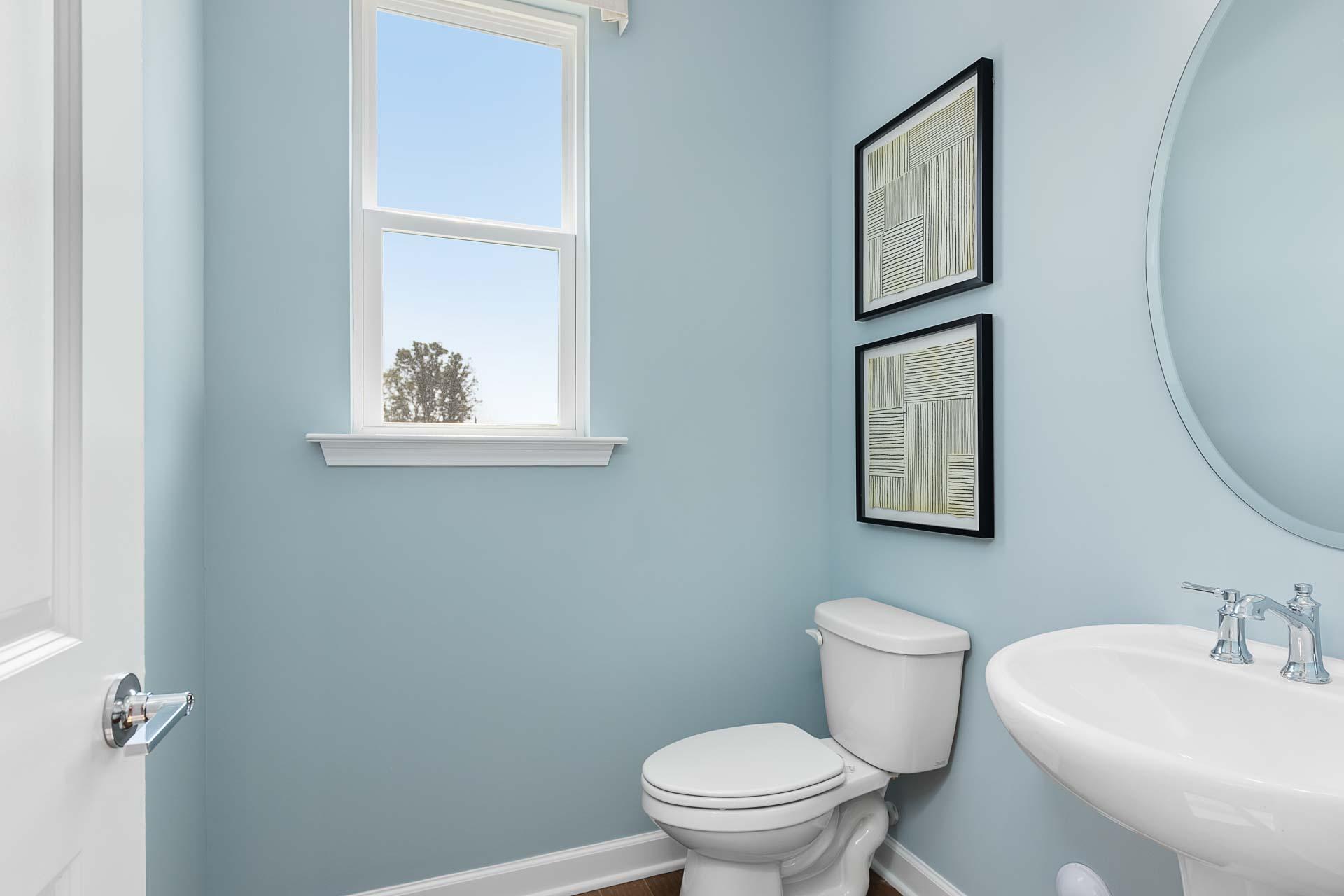 Light blue powder room at Highland Forest in Fuquay-Varina NC with white toilet, pedestal sink, round mirror and window