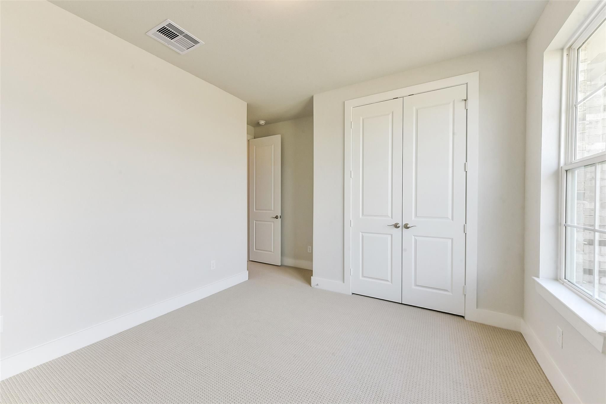Spacious empty bedroom with beige carpet, white walls, double closet doors, and large grid window in Davidson Homes The Edward A, Lago Mar, Texas City