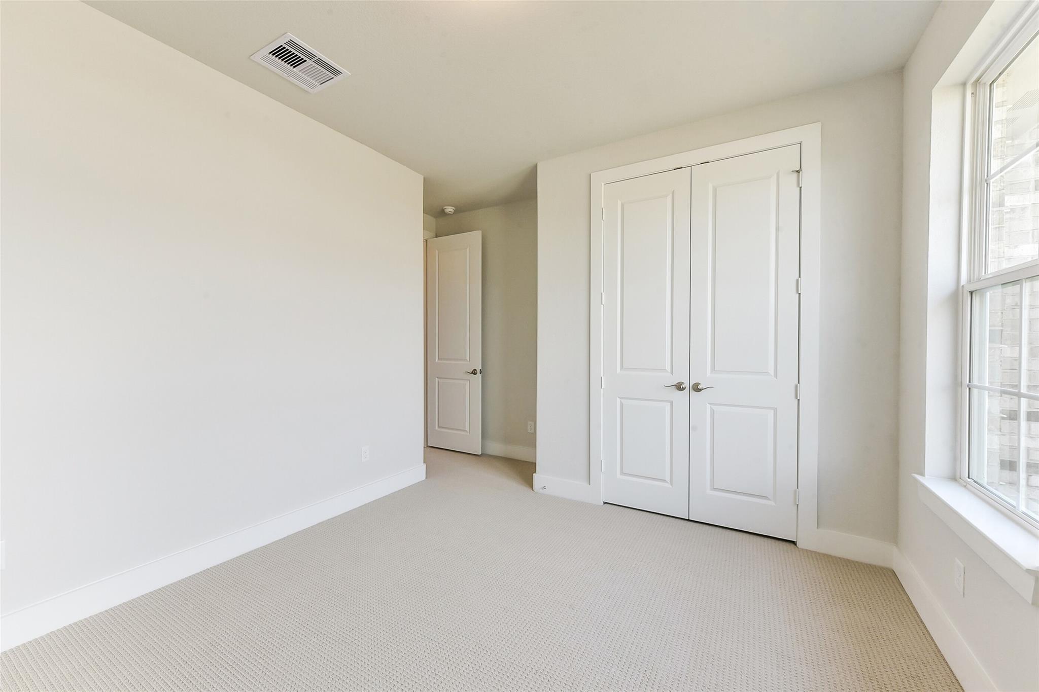 Spacious empty bedroom with beige carpet, white walls, double closet doors, and large grid window in Davidson Homes The Edward A, Lago Mar, Texas City