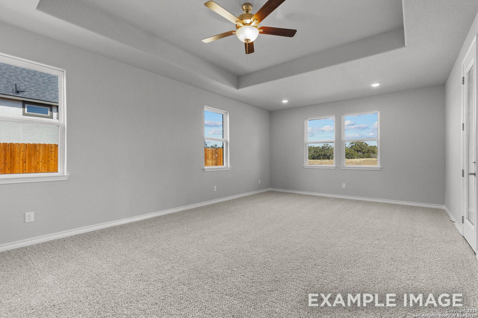 Spacious master bedroom in The Oxford H design with tray ceiling, neutral carpet, and large windows in Castroville, Texas.