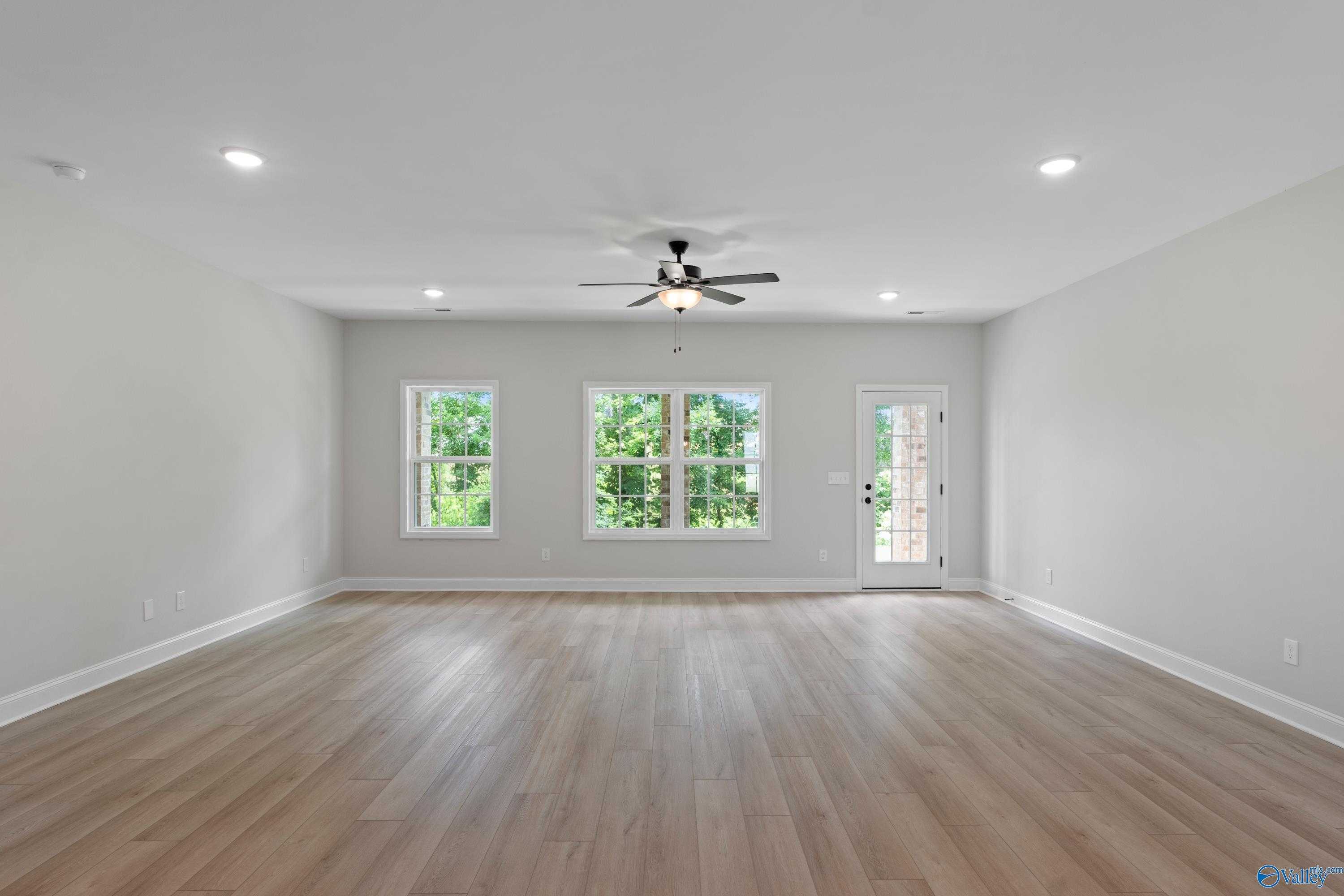 Spacious empty great room with hardwood floors, ceiling fan, large windows, and sliding door overlooking greenery in 3-bedroom Harvest AL home