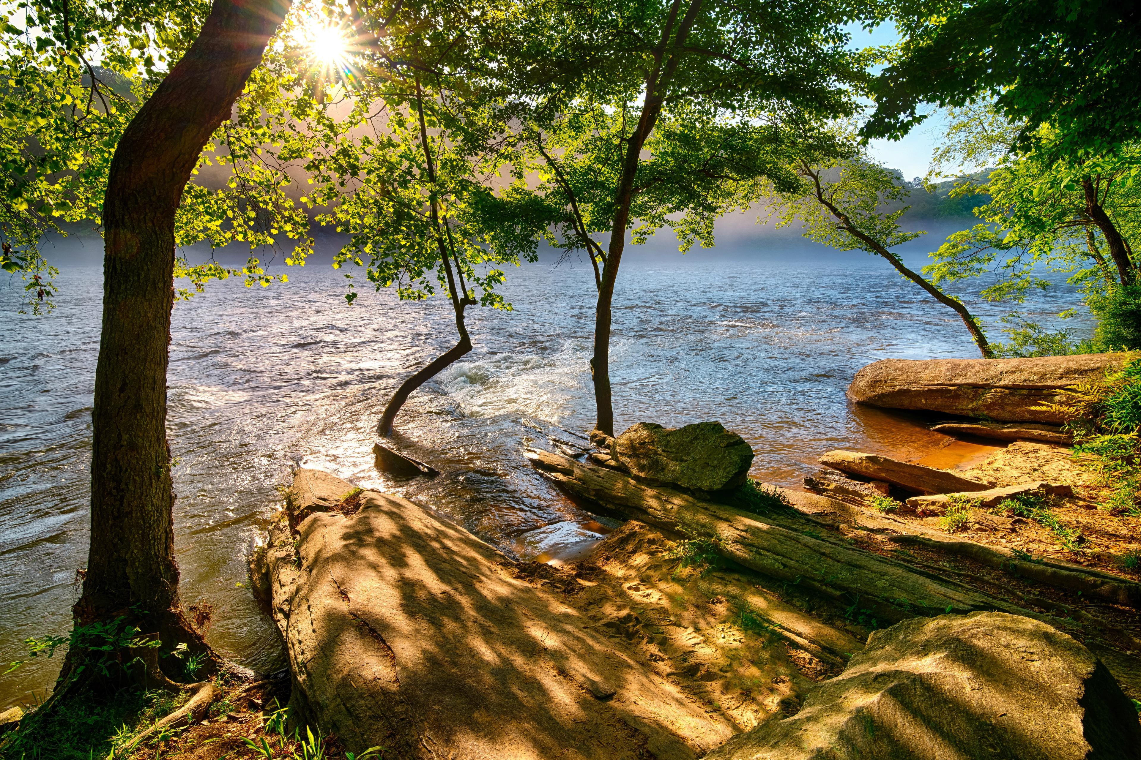Serene riverbank at The Village at Shallowford in Kennesaw GA with lush trees, sunlight rays, calm waters and rocks