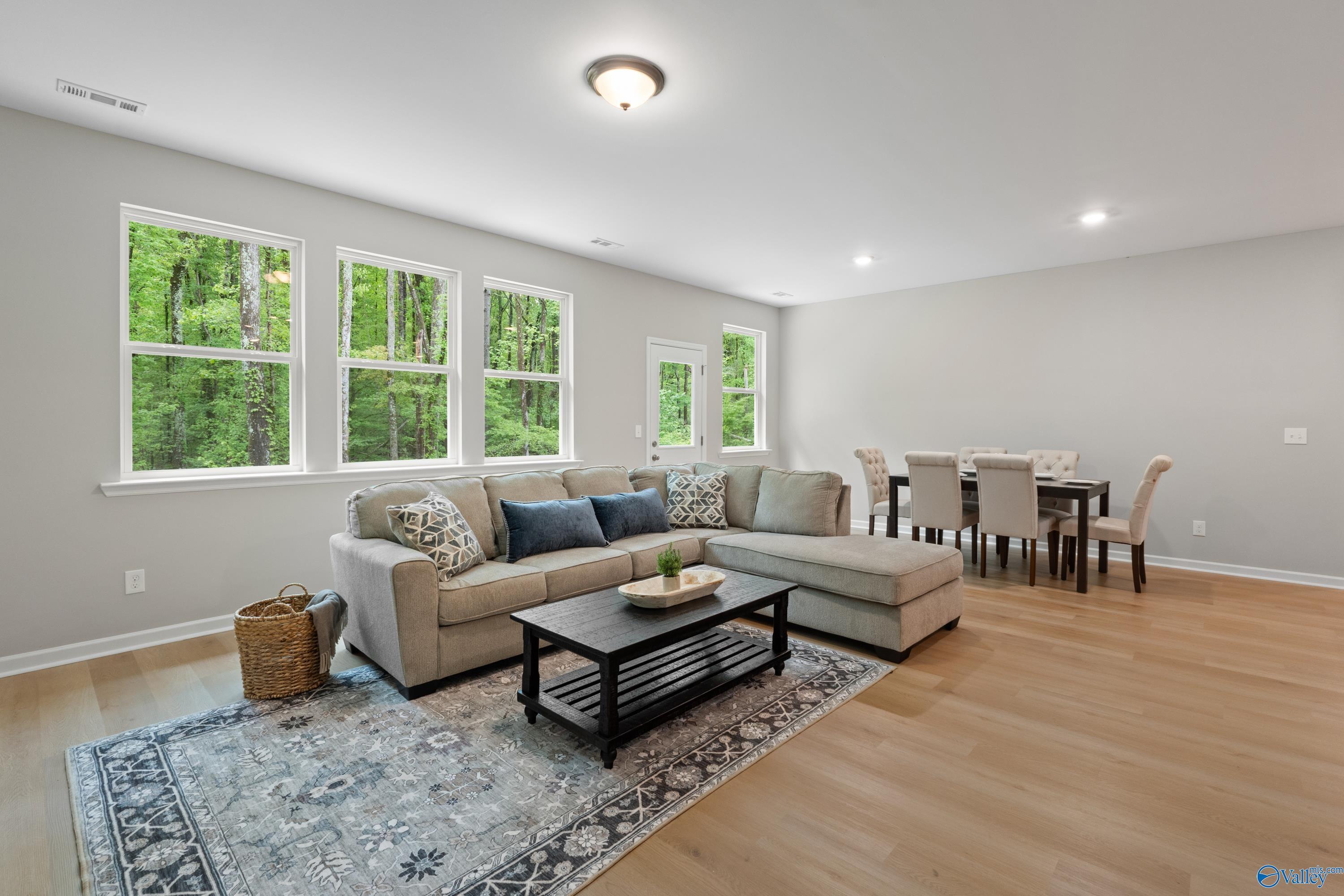 Open-concept living room with L-shaped sofa, dining table, hardwood floors, and large wooded windows in Davidson Homes The Luna, Hazel Green
