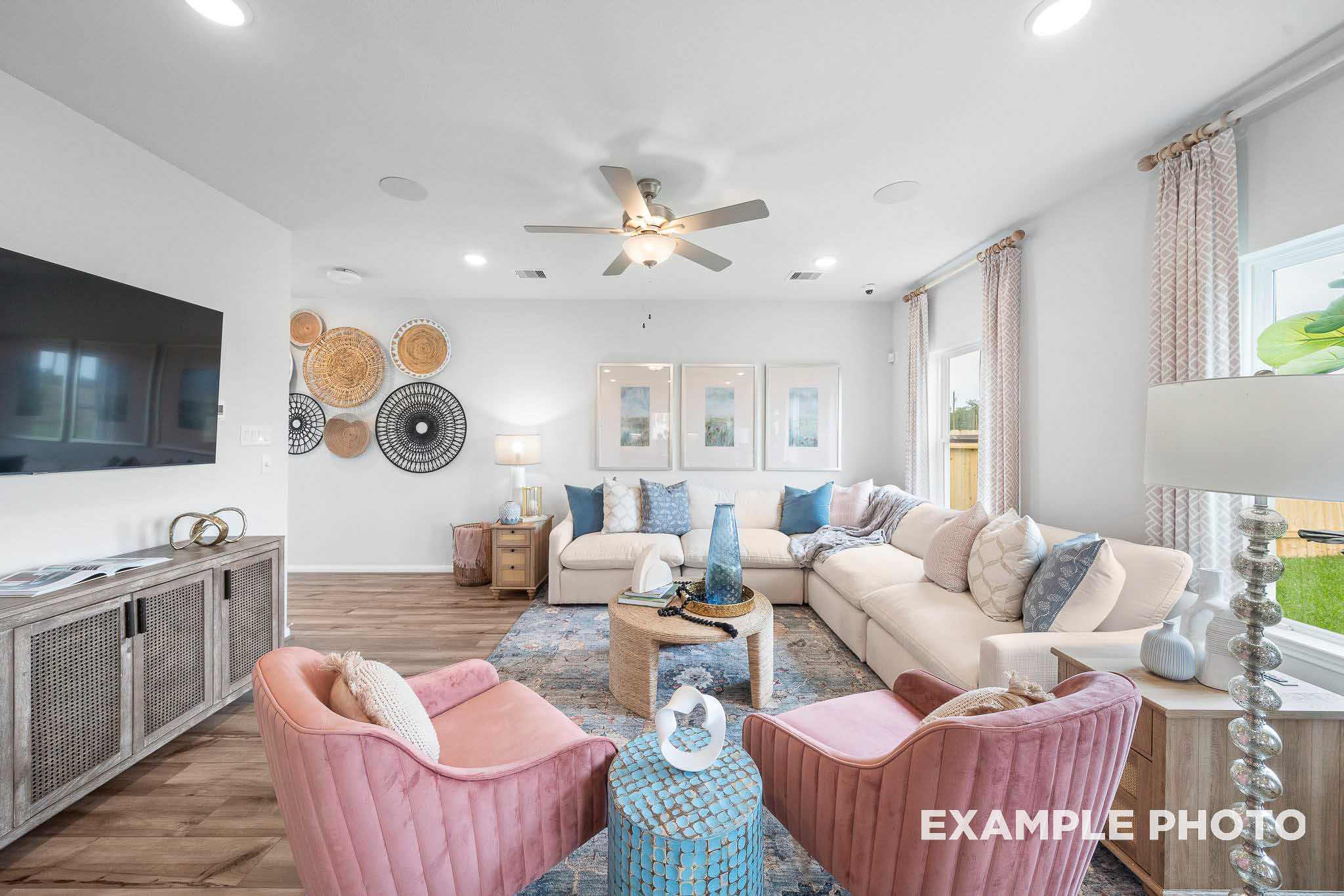 Spacious living room in The Trinity floor plan with beige sectional sofa, pink armchairs, woven wall hangings, and modern TV setup