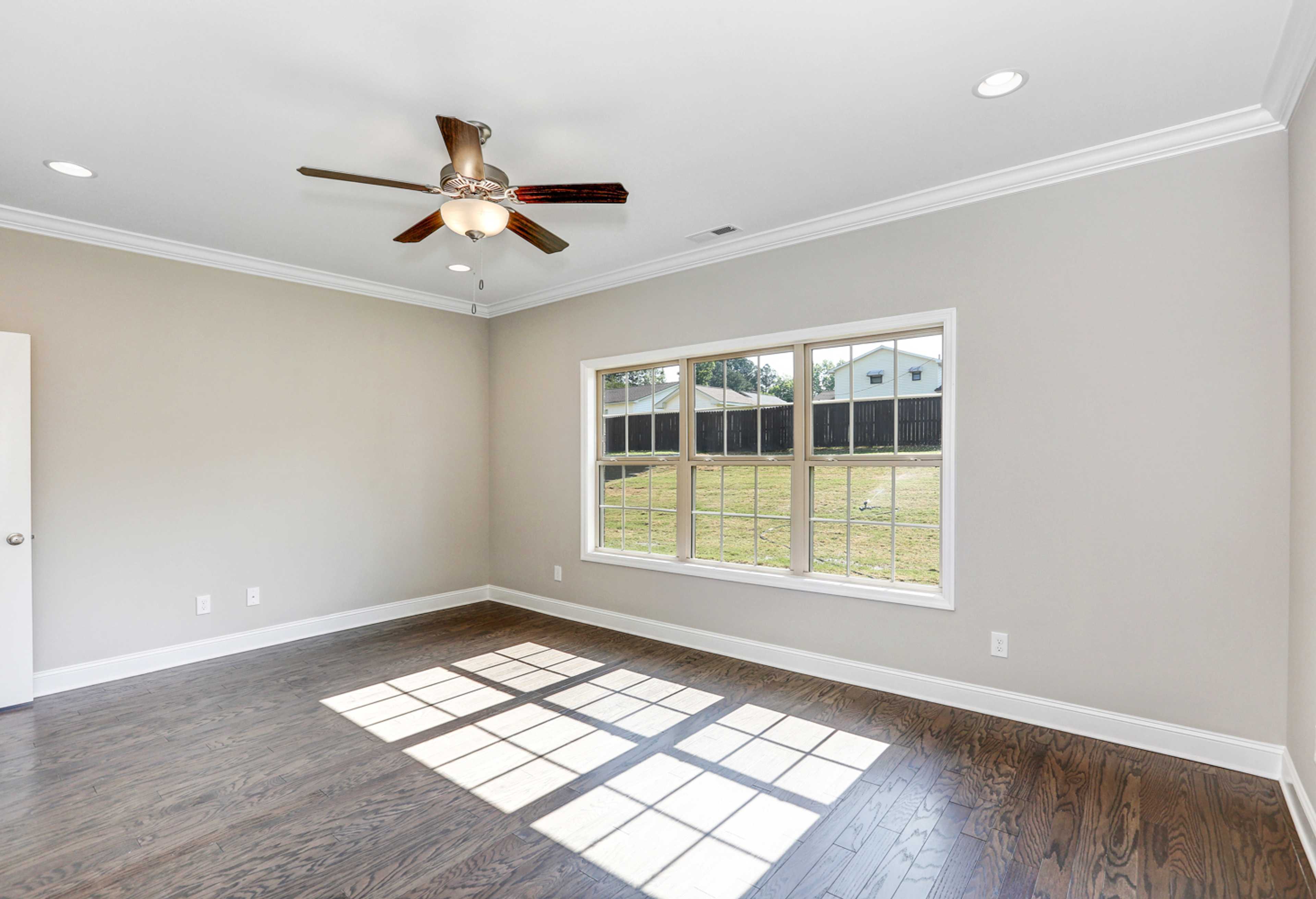 Spacious master bedroom in The Montgomery with light gray walls, hardwood floors, large triple windows, and ceiling fan