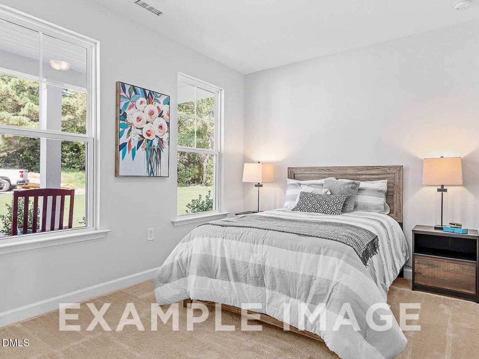 Bright secondary bedroom with queen bed, striped bedding, lamps, floral wall art, and yard views in Davidson Homes The Chestnut B, Lillington NC