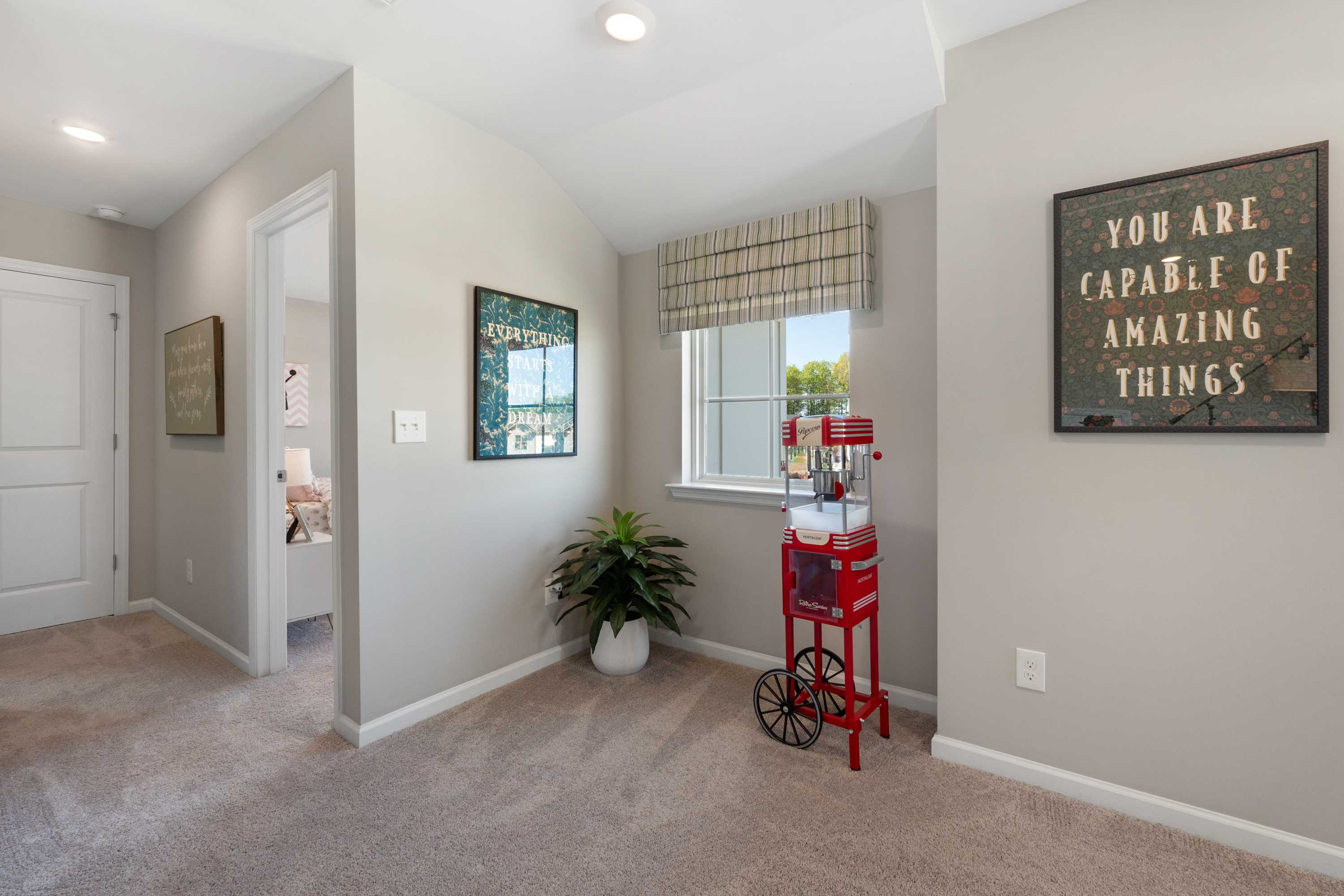 Spacious loft hallway in Evergreen Mill Madison AL home with beige walls, carpet floor, red vintage popcorn cart, potted plant, and inspirational wall art