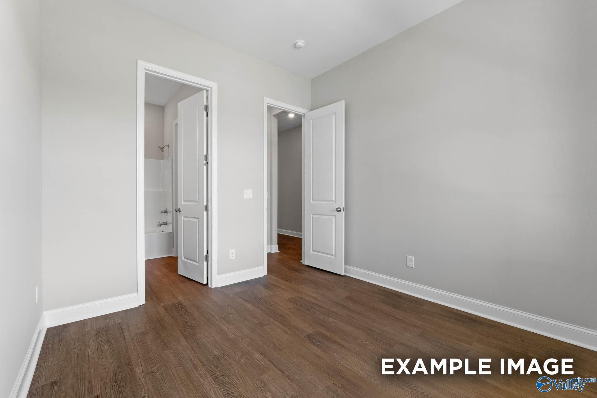 Spacious bedroom with light gray walls, hardwood floors, and open doors to dual bathrooms in Evermore Homes The Arcadia, Owens Cross Roads