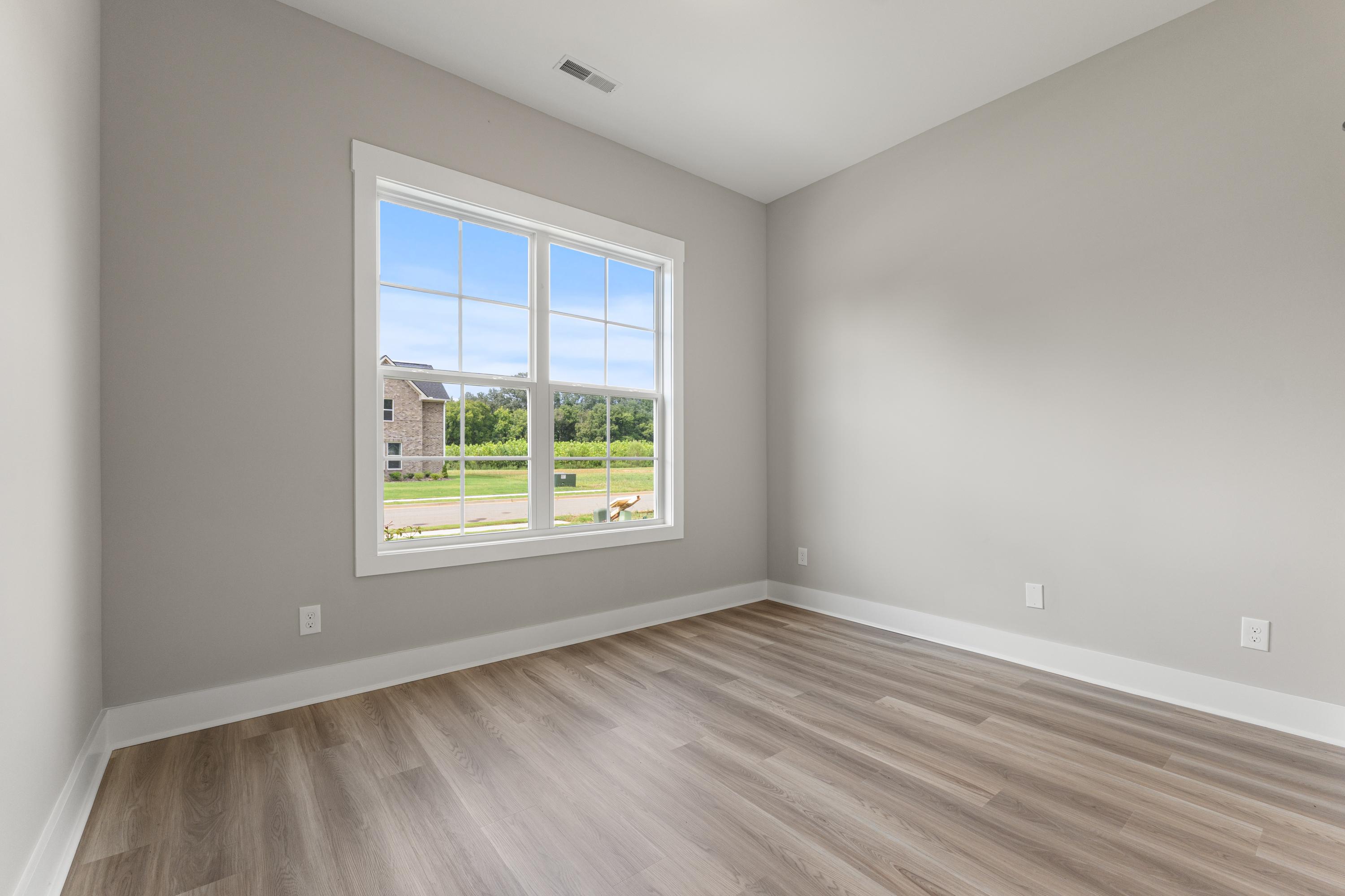 Spacious bedroom in The Oxford home plan with light gray walls, wood laminate floors, and large window overlooking green lawn