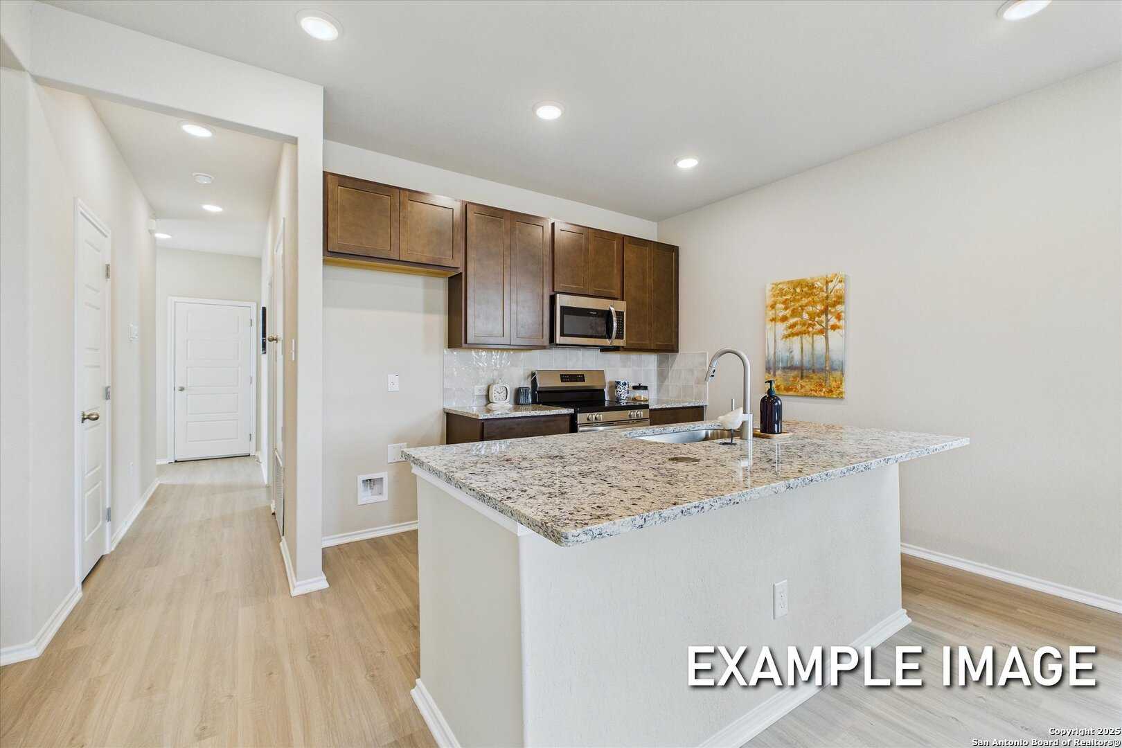 Modern kitchen with granite island, stainless appliances, brown cabinets, and open hallway in Davidson Homes The Comal B, Agave, San Antonio