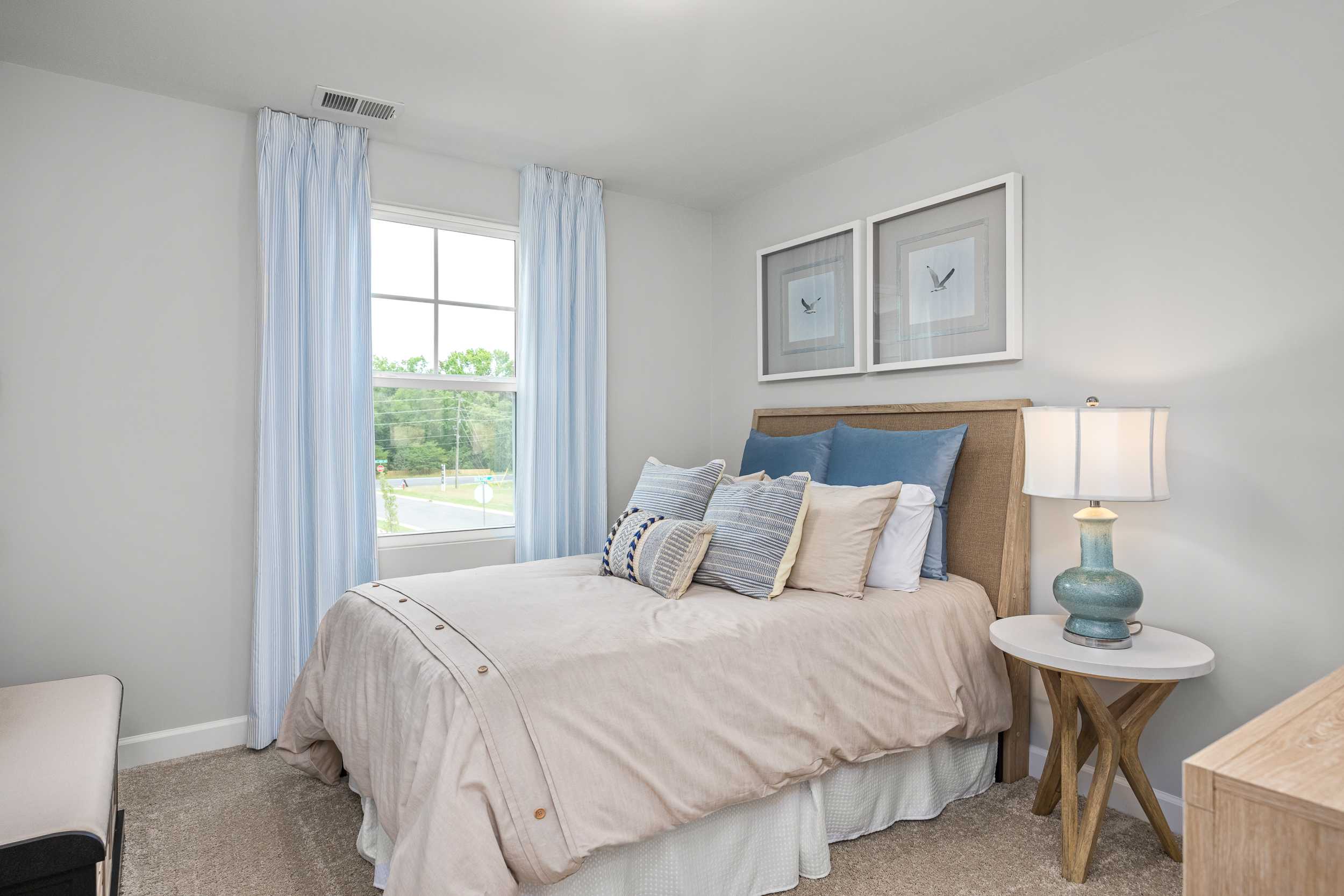Cozy bedroom at Springvale in Fuquay-Varina NC with queen bed, patterned pillows, bedside lamp, and large window with blue curtains