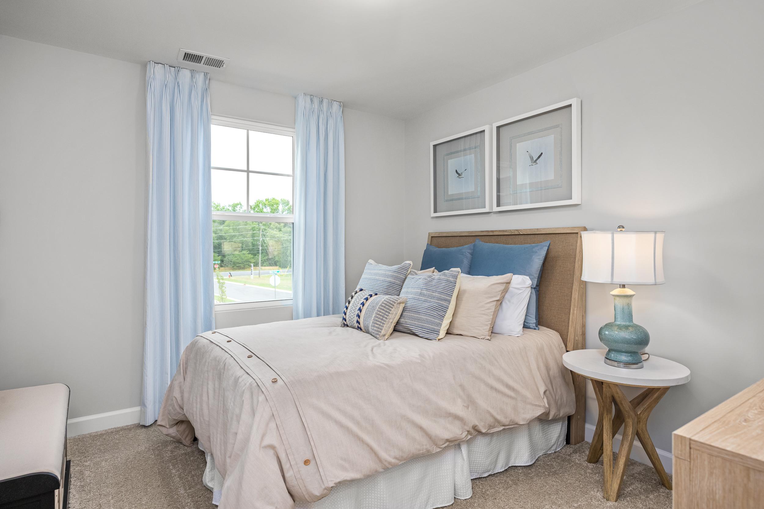 Cozy bedroom at Springvale in Fuquay-Varina NC with queen bed, patterned pillows, bedside lamp, and large window with blue curtains