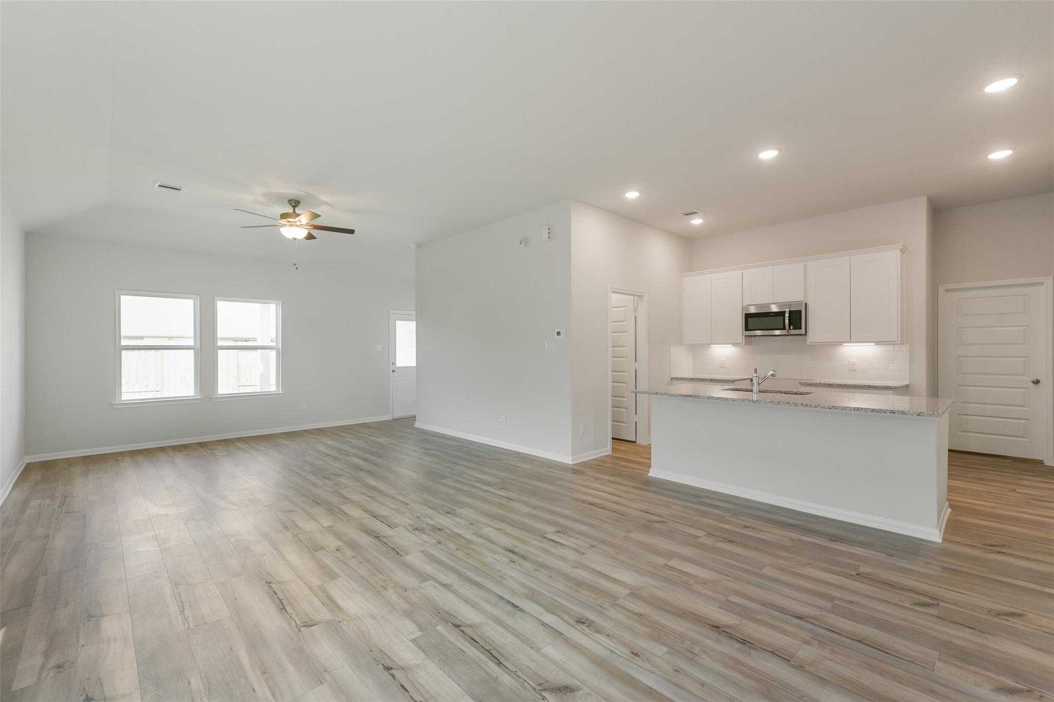 Open-concept living and kitchen area with white cabinetry, wood laminate floors, ceiling fan, and large windows in Davidson Homes The Laguna B, Dayton, Texas