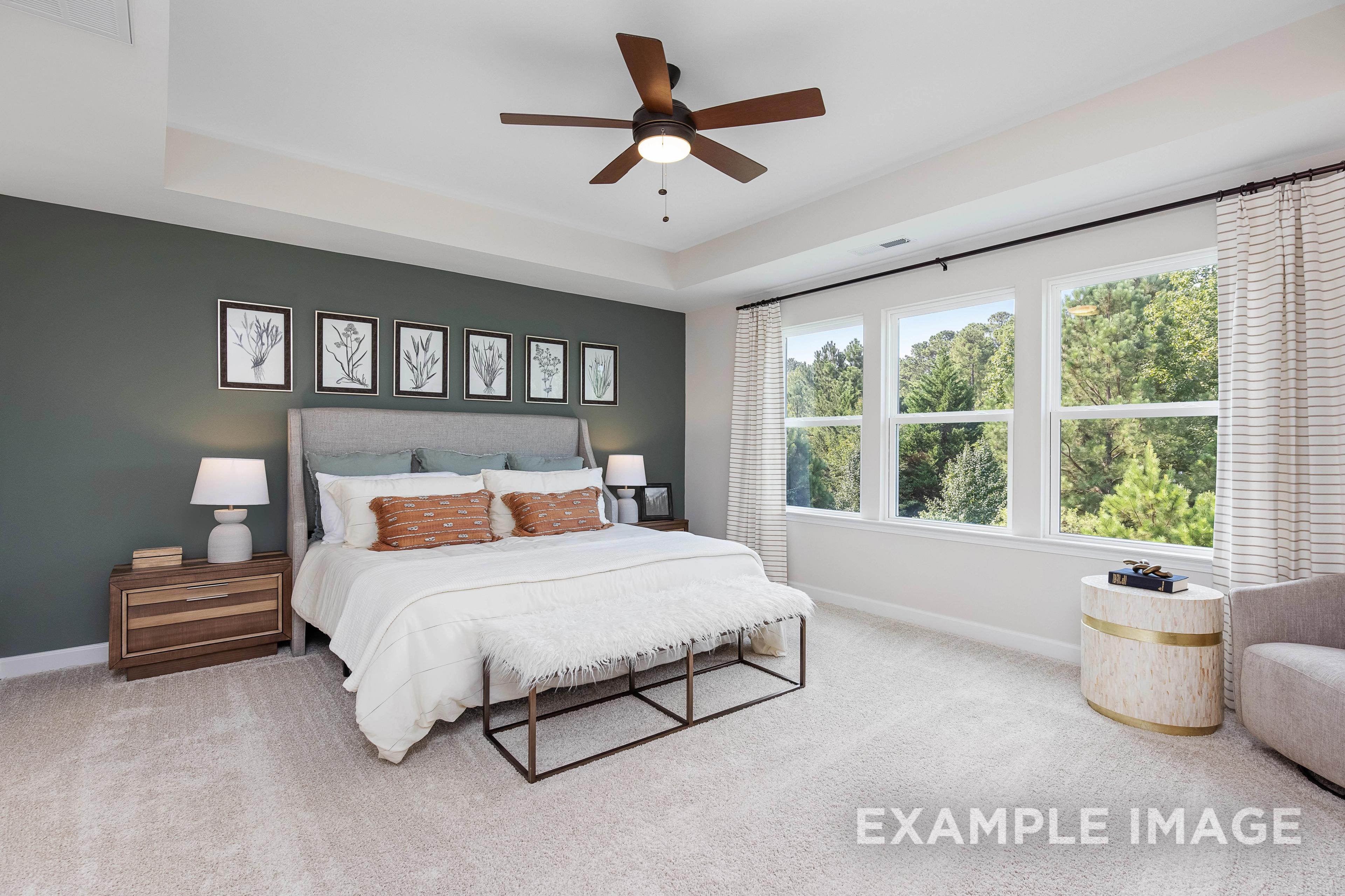 Spacious master bedroom in The Willow B with gray accent wall, king bed, large windows overlooking woods, ceiling fan
