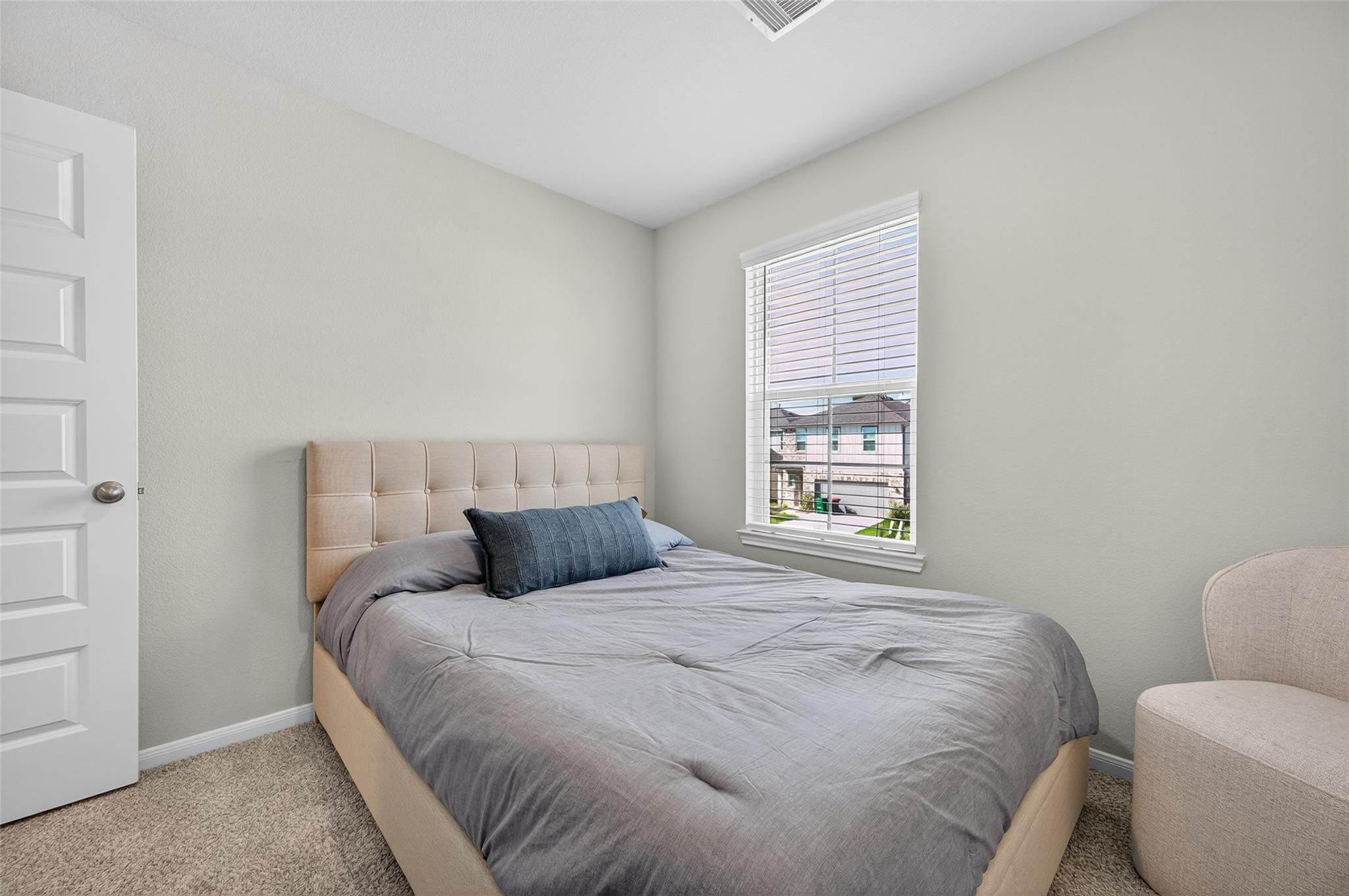 Cozy secondary bedroom featuring neutral gray walls, queen bed with gray bedding, window view, and armchair in Davidson Homes The Brazos E, Magnolia, Texas