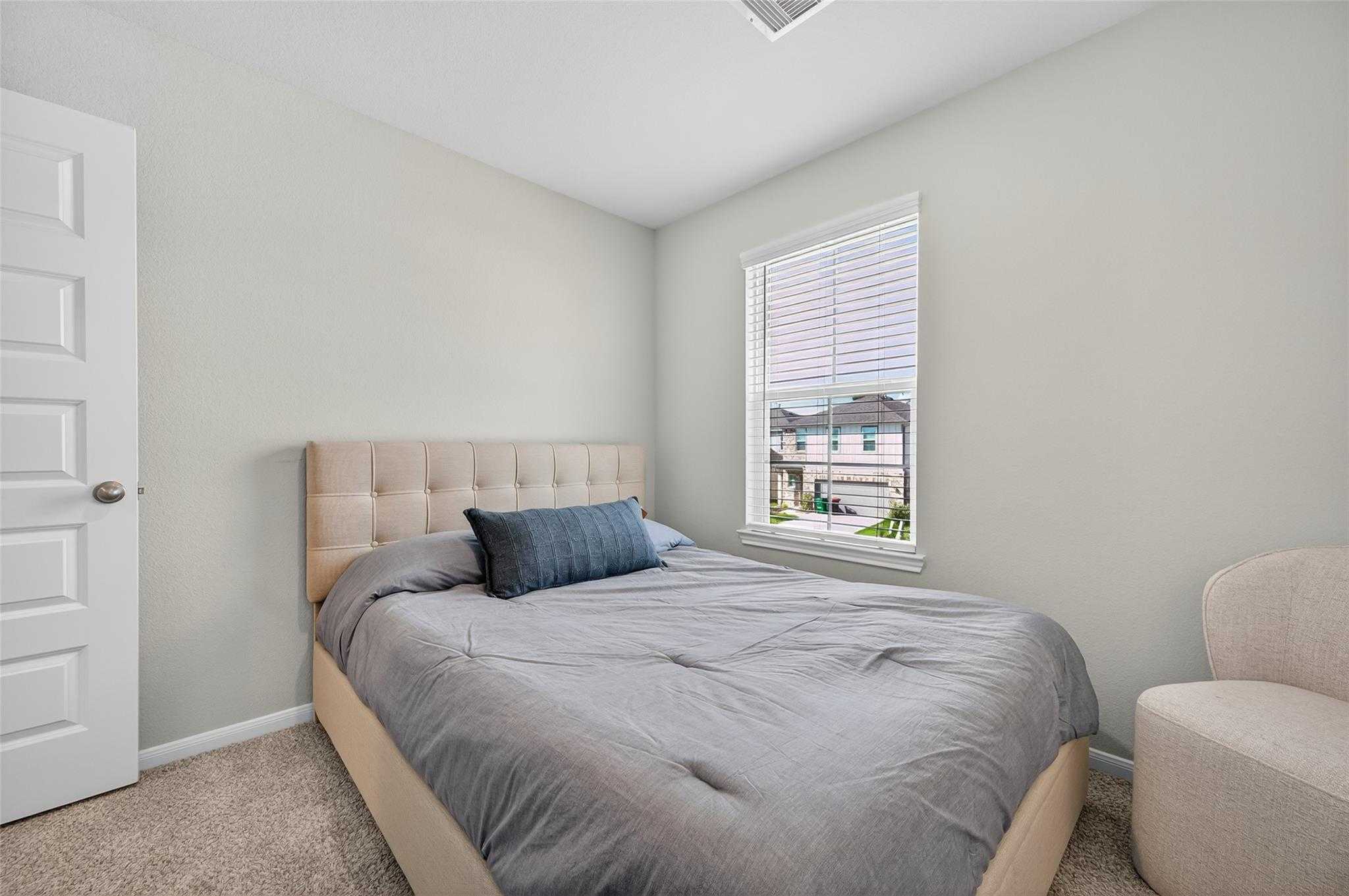 Cozy secondary bedroom featuring neutral gray walls, queen bed with gray bedding, window view, and armchair in Davidson Homes The Brazos E, Magnolia, Texas