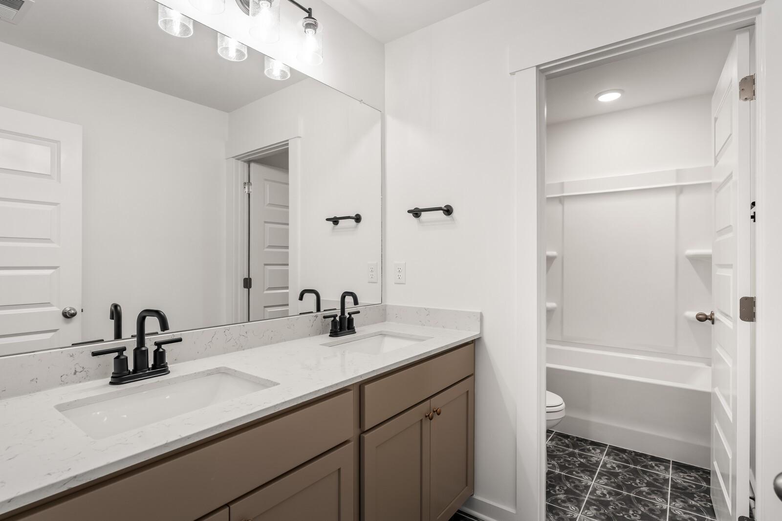 Modern double vanity with black faucets, quartz counters, brown cabinets, and walk-in shower in Davidson Homes The Ash A, Gallatin, TN