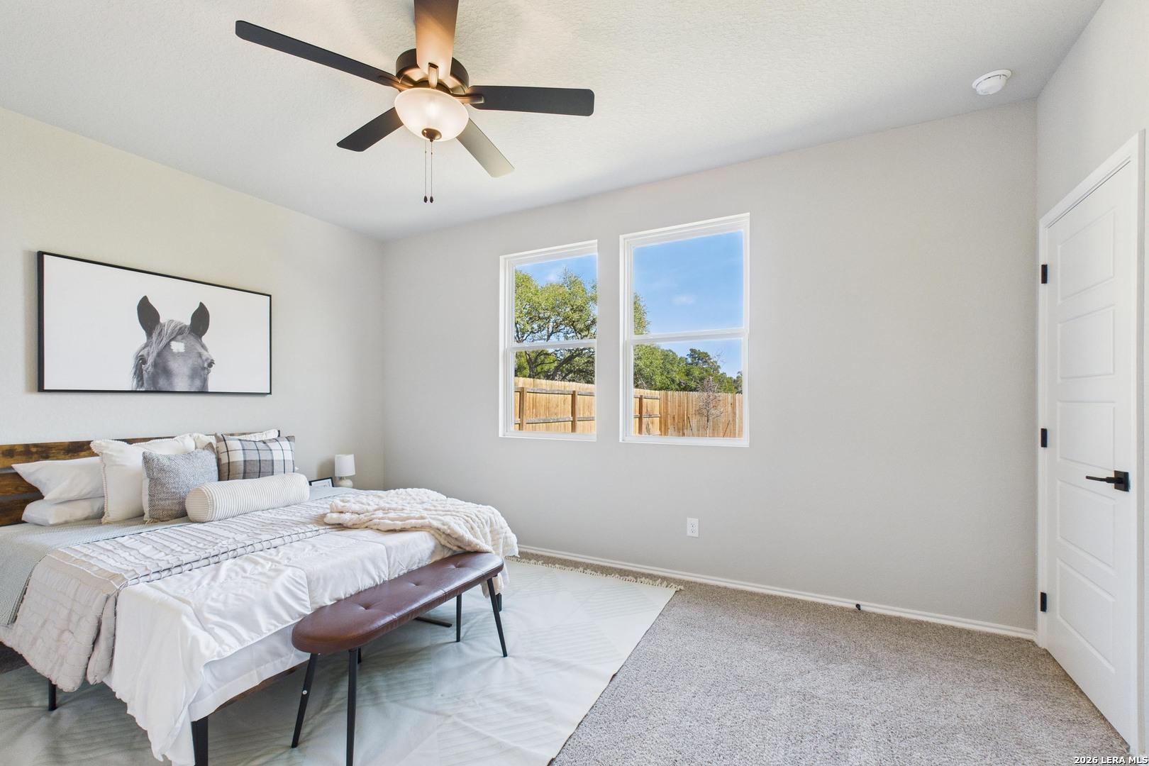 Cozy bedroom with white bedding on wooden bed frame, ceiling fan, large windows overlooking backyard fence and trees, horse artwork in Davidson Homes The Douglas E, San Antonio
