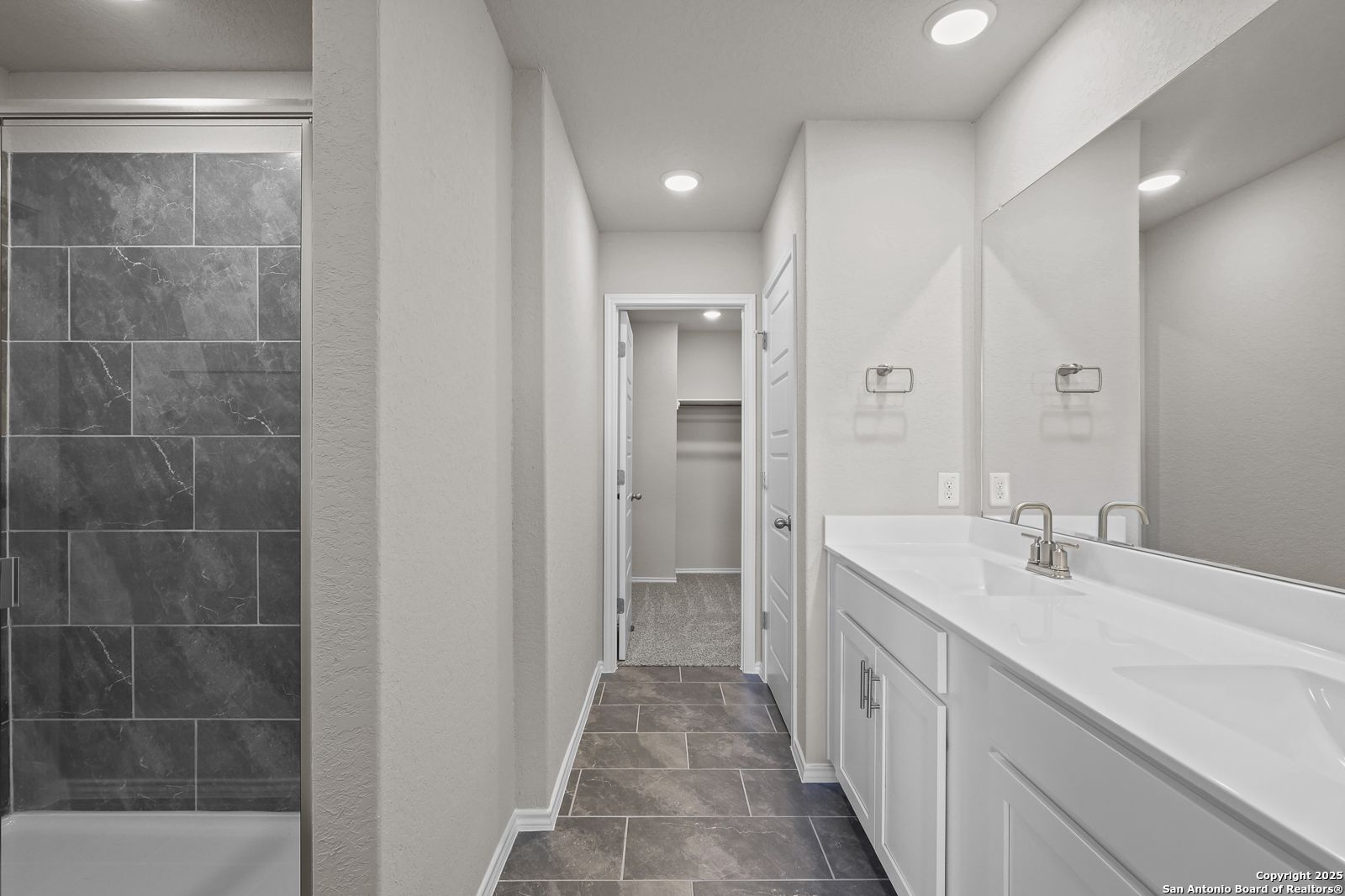 Modern master bathroom with slate-tile walk-in shower, double white vanity, mirrors, and closet access in Davidson Homes The Trinity D, San Antonio