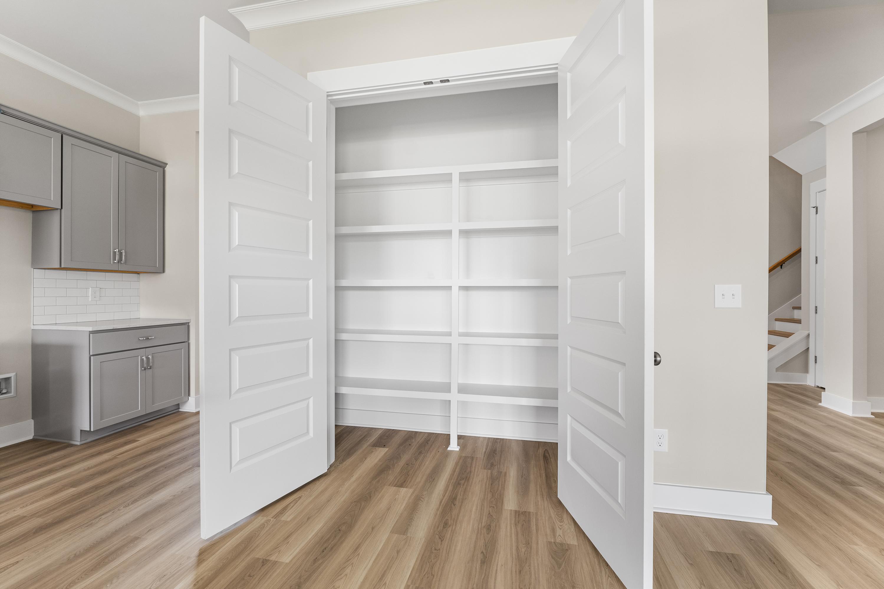 Spacious walk-in pantry in The Oxford A with open white doors and tiered shelves, adjacent gray cabinet kitchen and beige walls