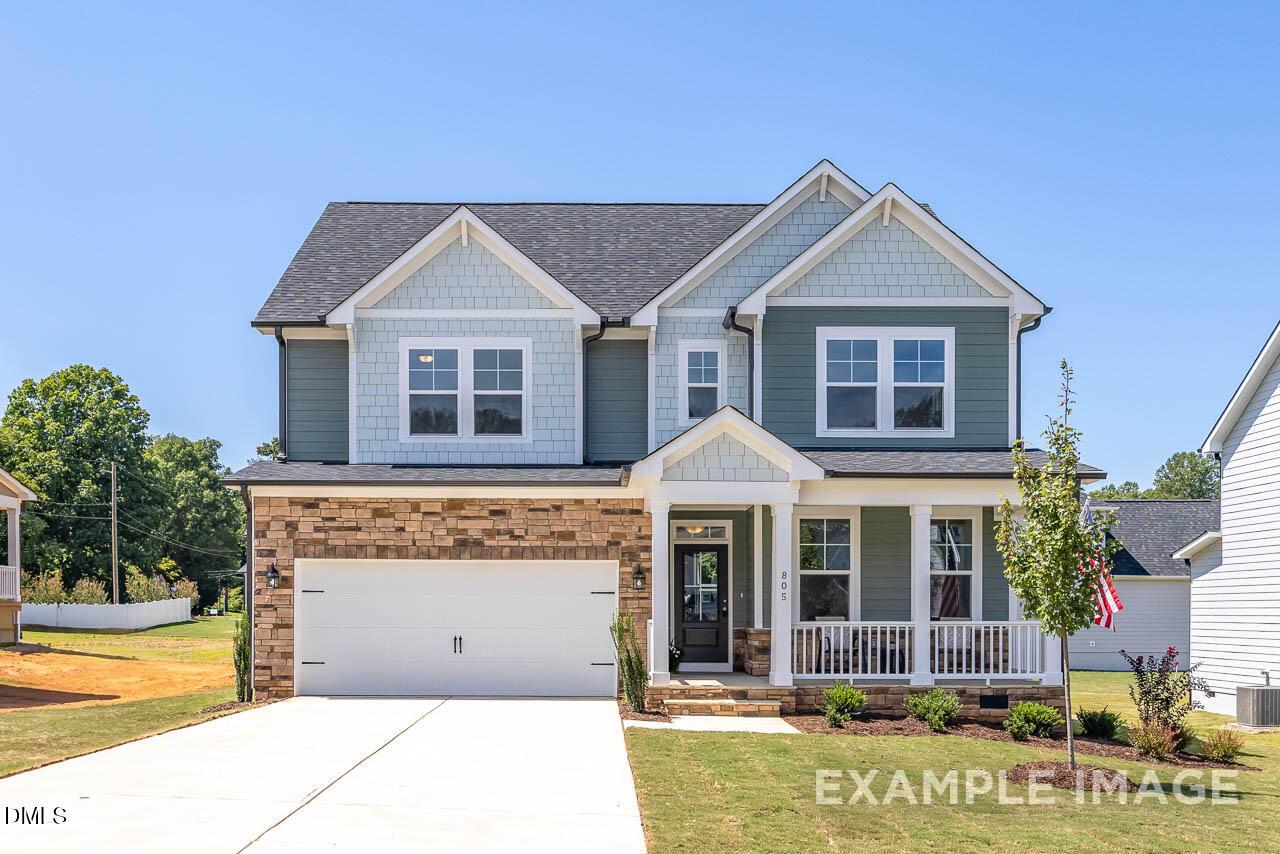Two-story light blue Craftsman home with brick 2-car garage, covered porch, and flag in Retreat at North Main, Lillington, NC